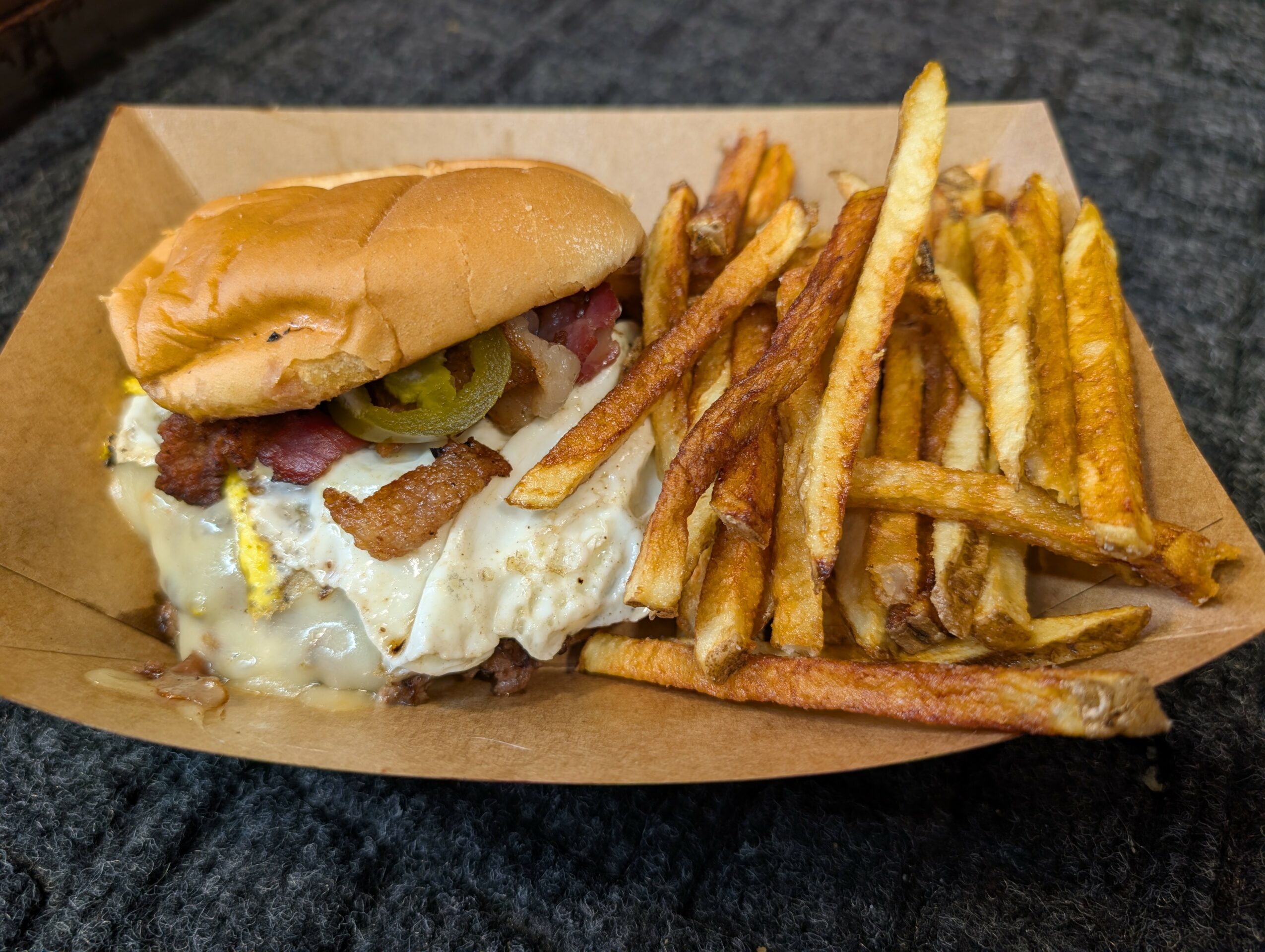 Sandwich with fried egg, bacon, pickles, and bread, served with French fries on a tray.
