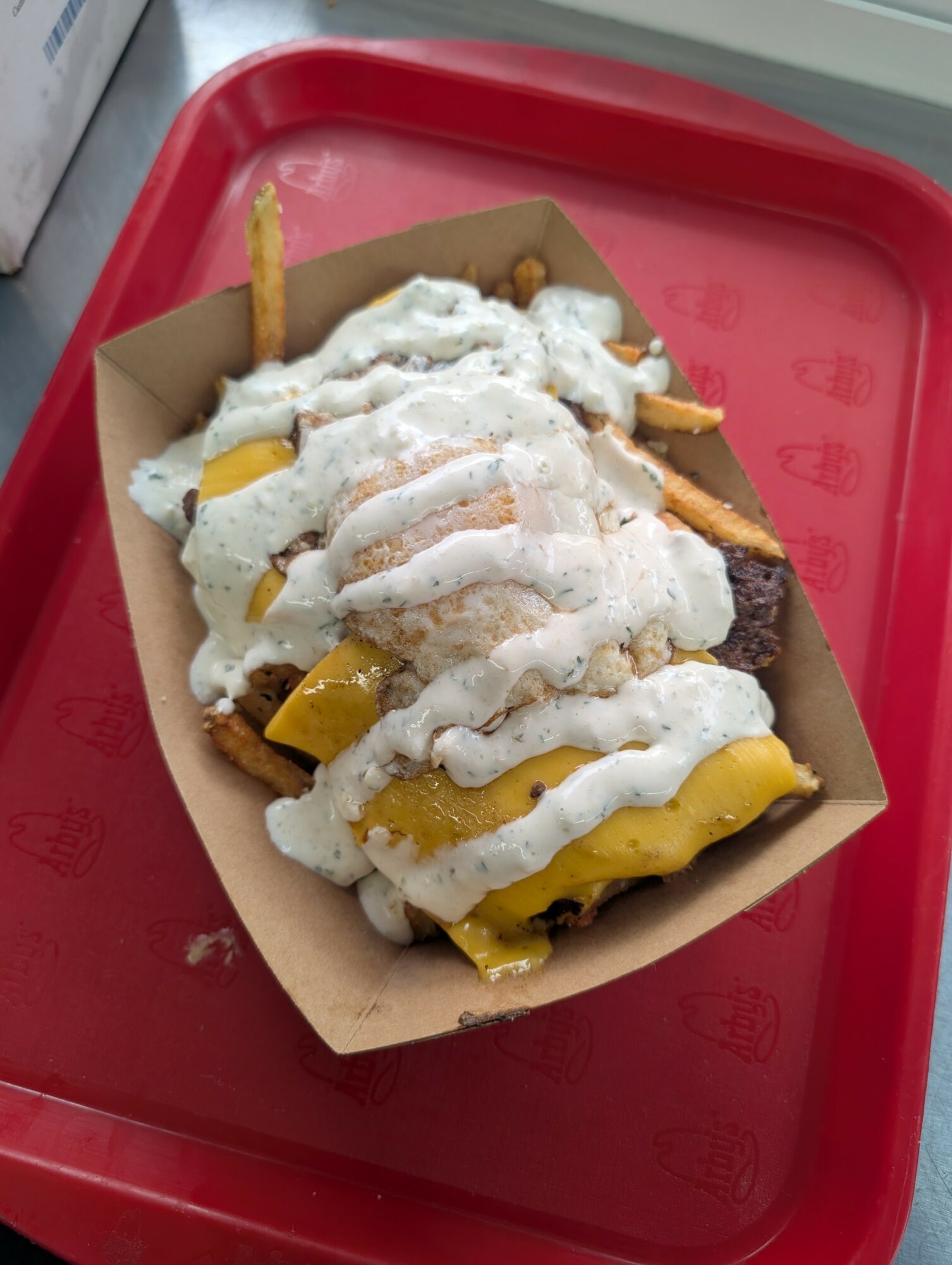 French fries topped with melted cheese and white sauce on a paper tray, placed on a red tray.