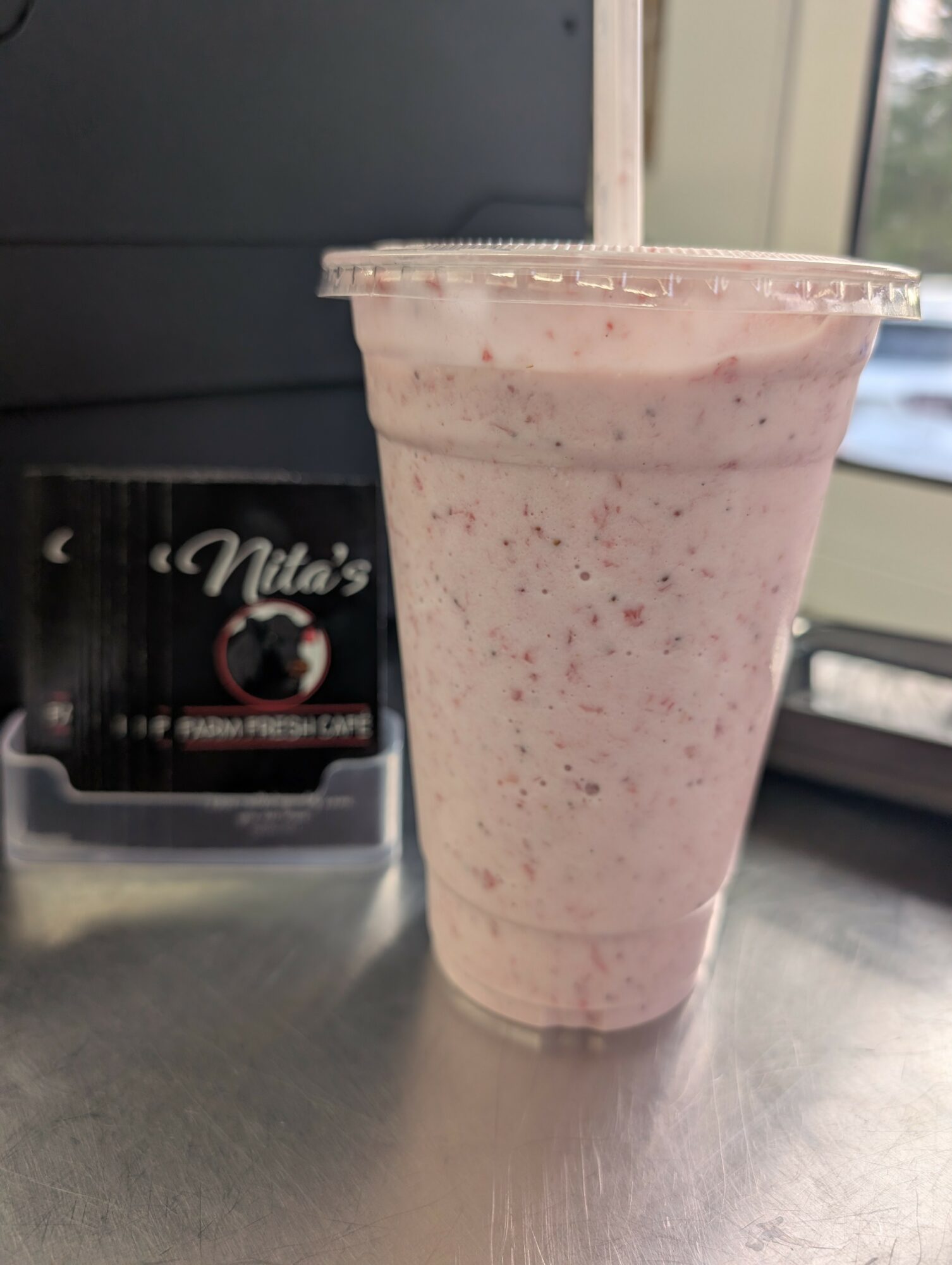 A pink speckled smoothie in a clear plastic cup on a metallic surface, with a Nita's coffee packet in the background.