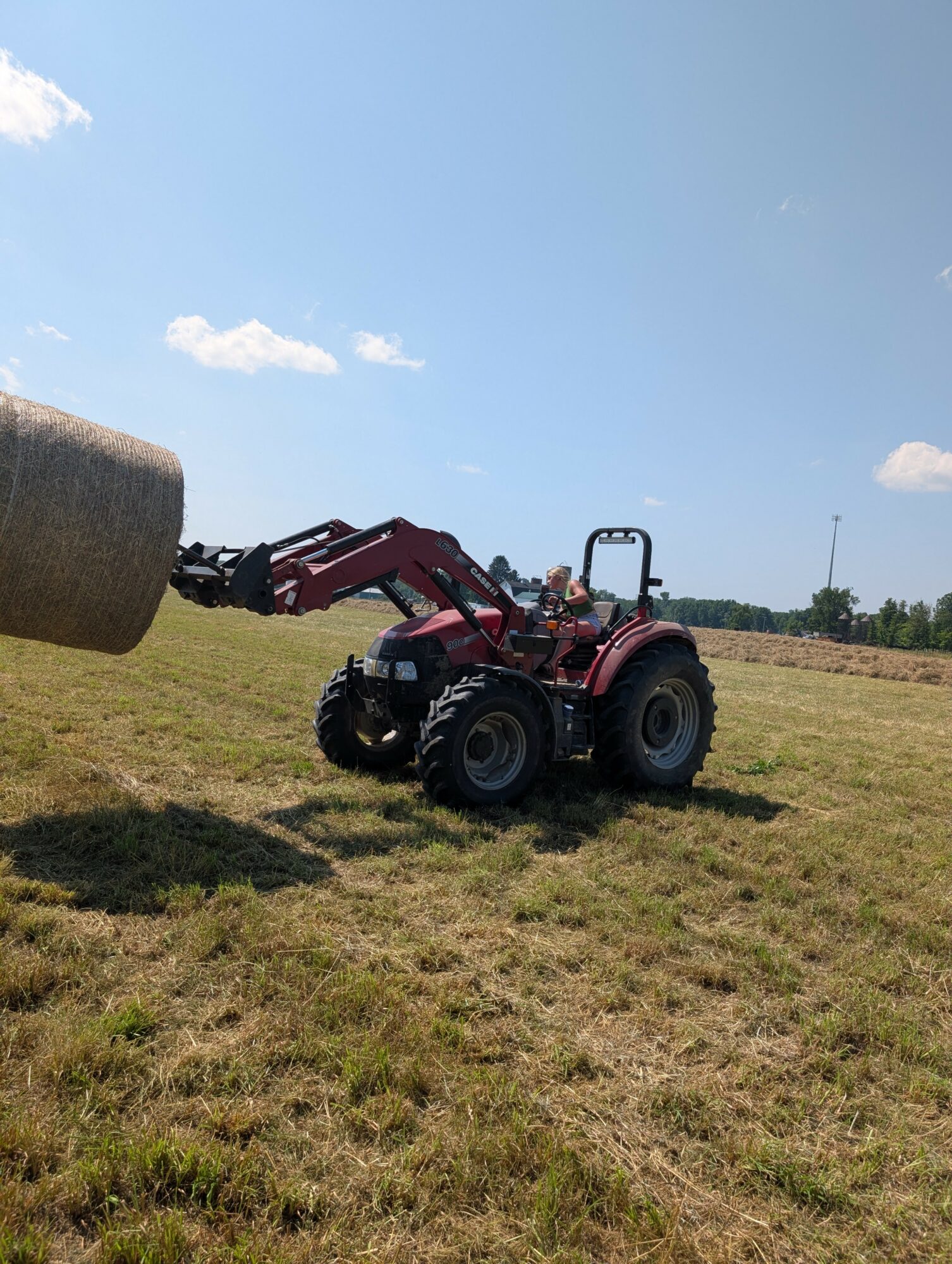 Red tractor lifting a large hay bale in an open field under a blue sky with clouds.
