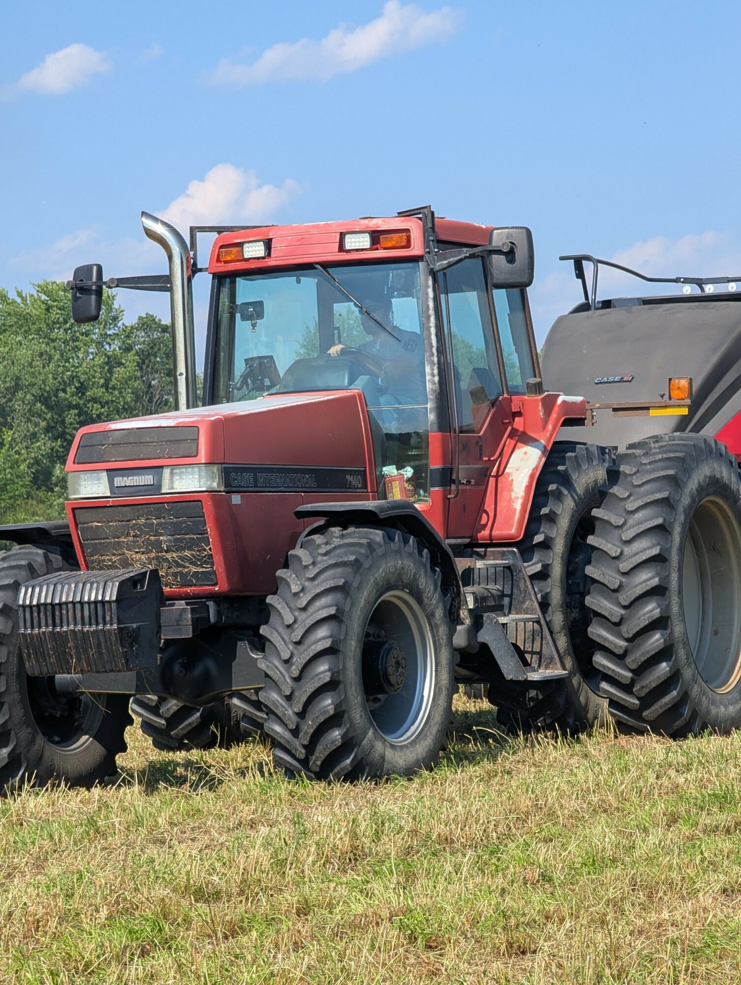 Red tractor with large tires on grassy field under blue sky with clouds