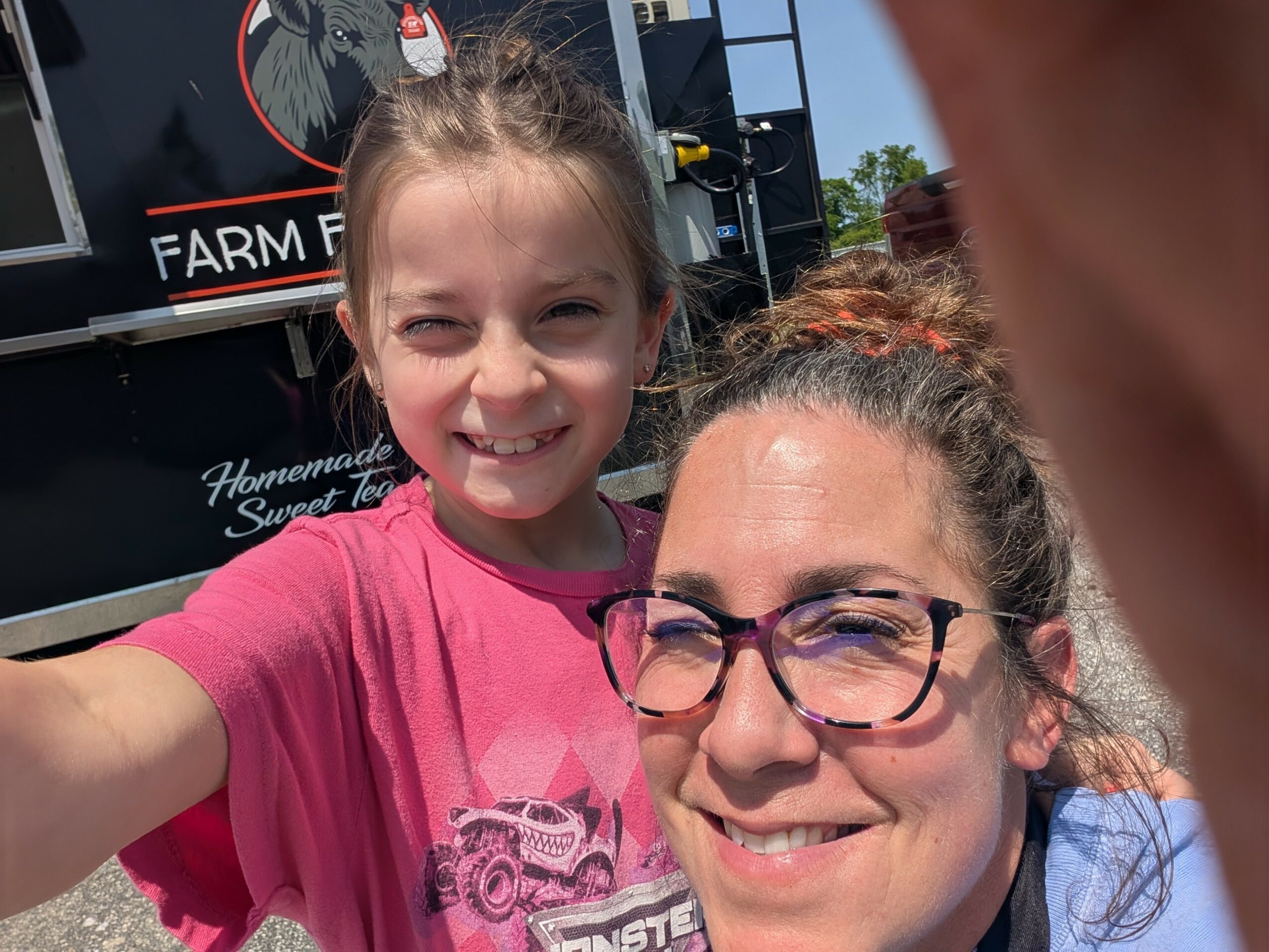 Two smiling girls outdoors, one wearing glasses and a pink shirt, the other in a pink shirt, with a farm sign in background.