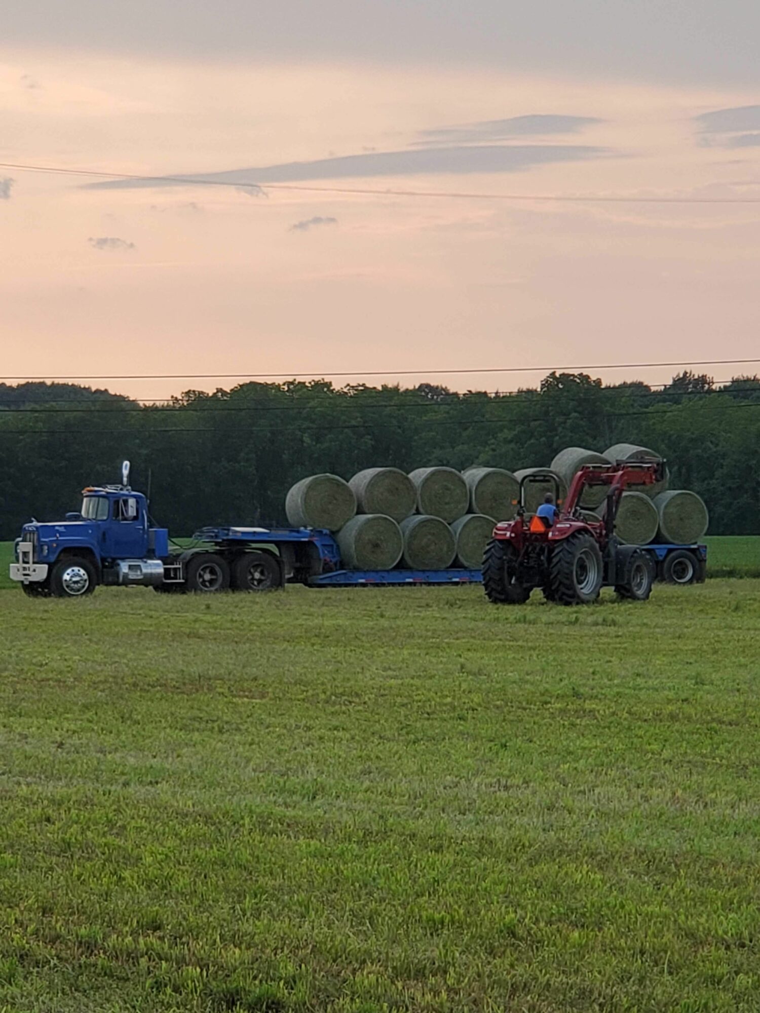 A tractor with a trailer is parked on a grassy field during sunset, with trees in the background.