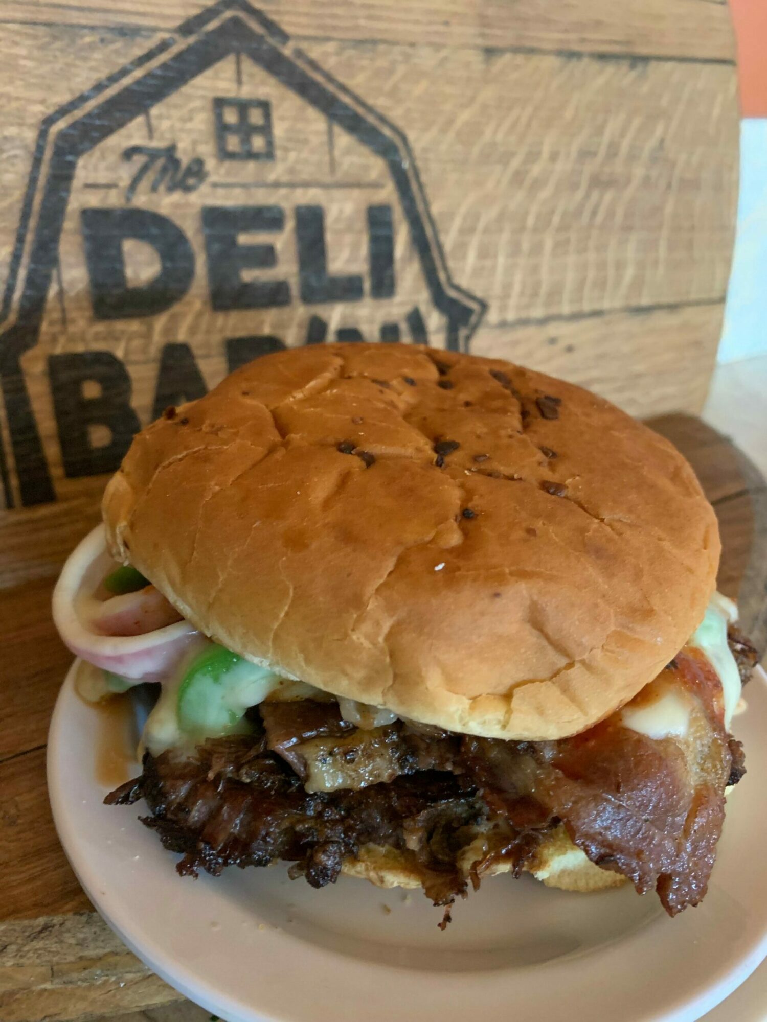 Sandwich with meat, cheese, and vegetables in a bun on a white plate, on a wooden surface, with a logo partially visible.