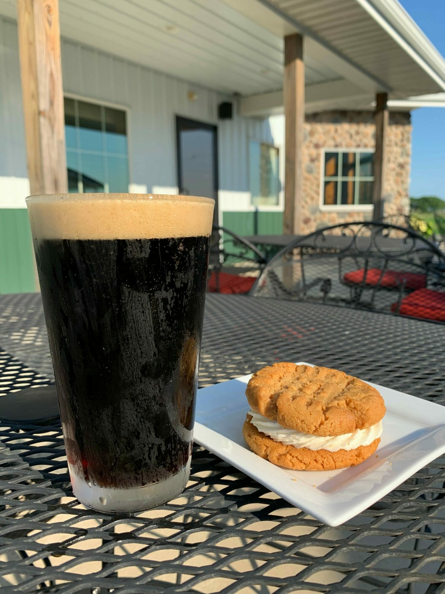A glass of dark soda with a straw and a cookie on a plate on a metal outdoor table.