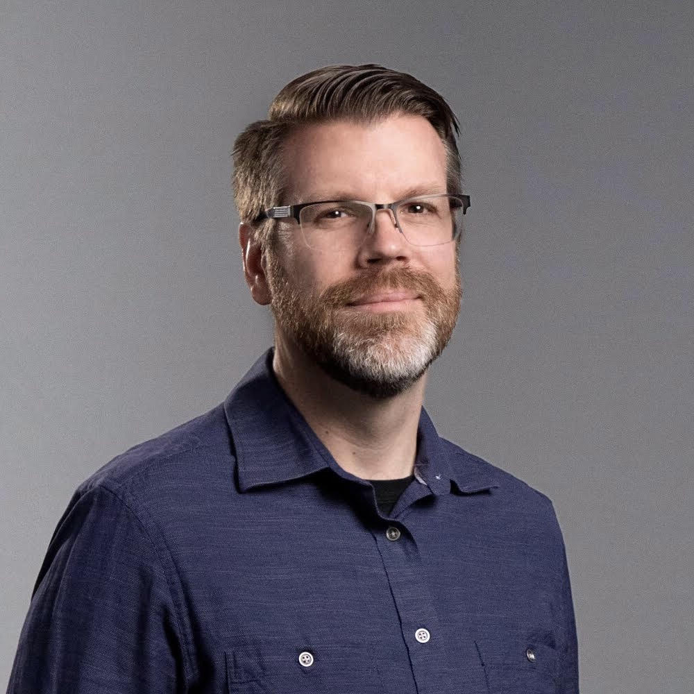 Man with glasses, beard, and short hair wearing a dark blue shirt, looking at the camera against a gray background.