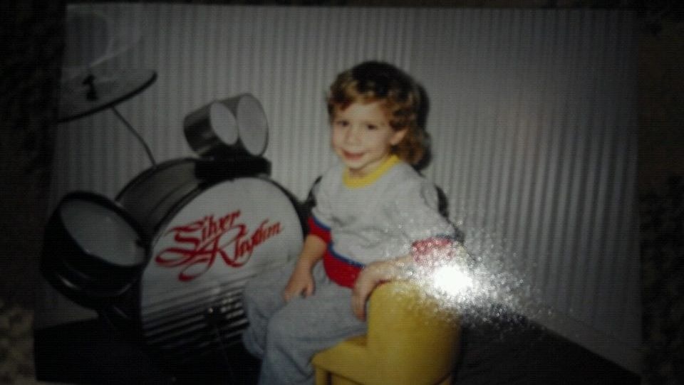 Child with curly hair sitting on a yellow chair, smiling, with a robot-like object nearby, against a striped background.