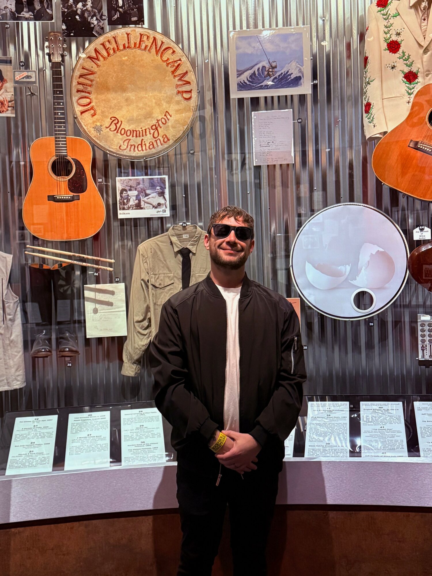 Young man with sunglasses smiling in front of a display with guitars, a sign, and framed pictures.