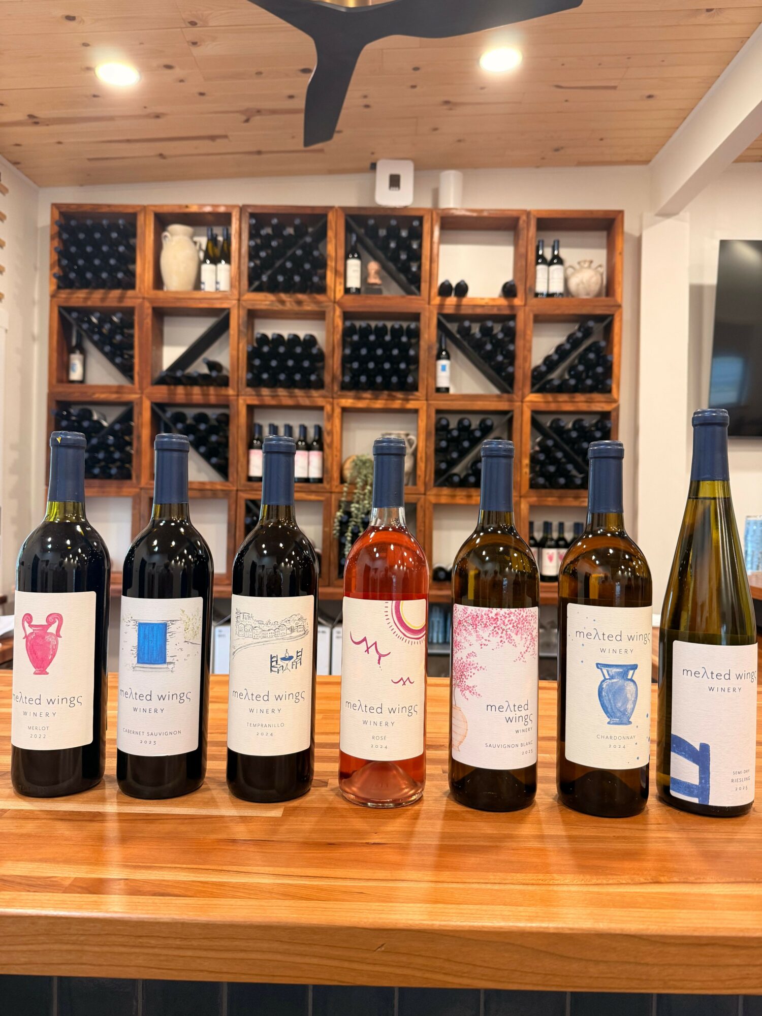 Display of eight wine bottles on a wooden table with a wine rack in the background.