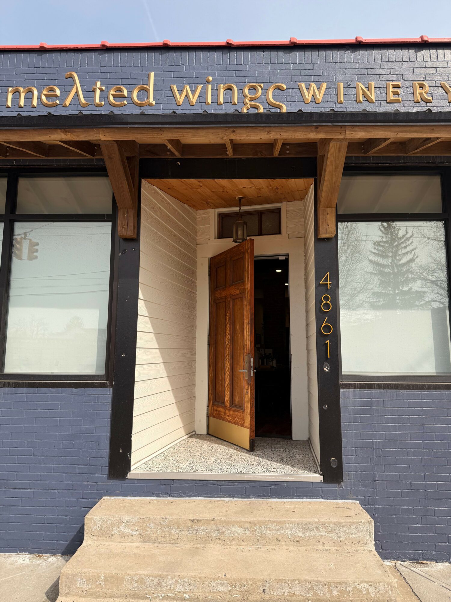 Open front door with steps leading to entrance, modern building exterior with large windows, wooden accents, and a blue wall.