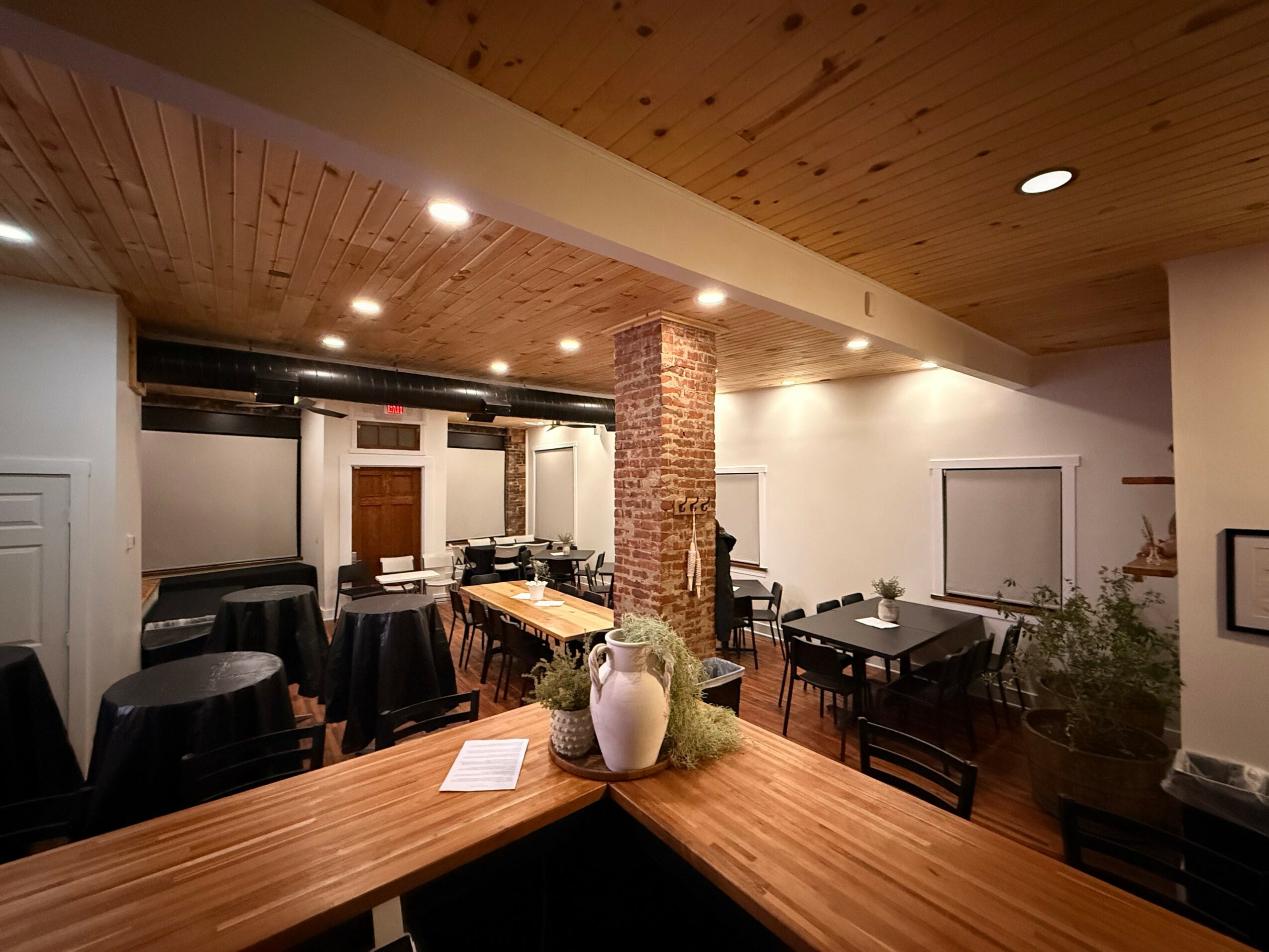 Interior of a restaurant with wooden ceiling, brick column, tables, and chairs, decorated with vases and plants.