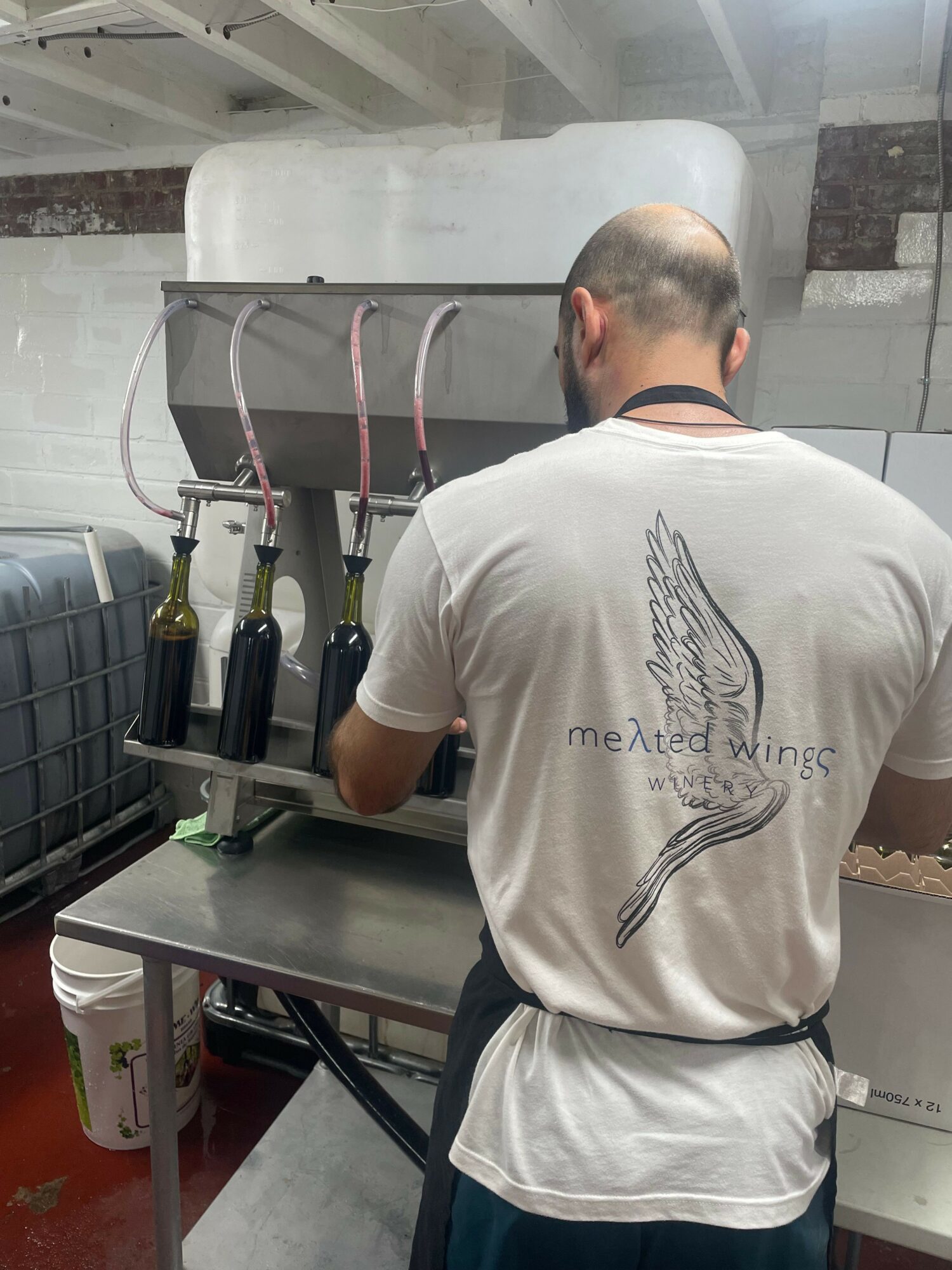 Person with short hair wearing a white t-shirt with a wing design, working at a stainless steel counter.