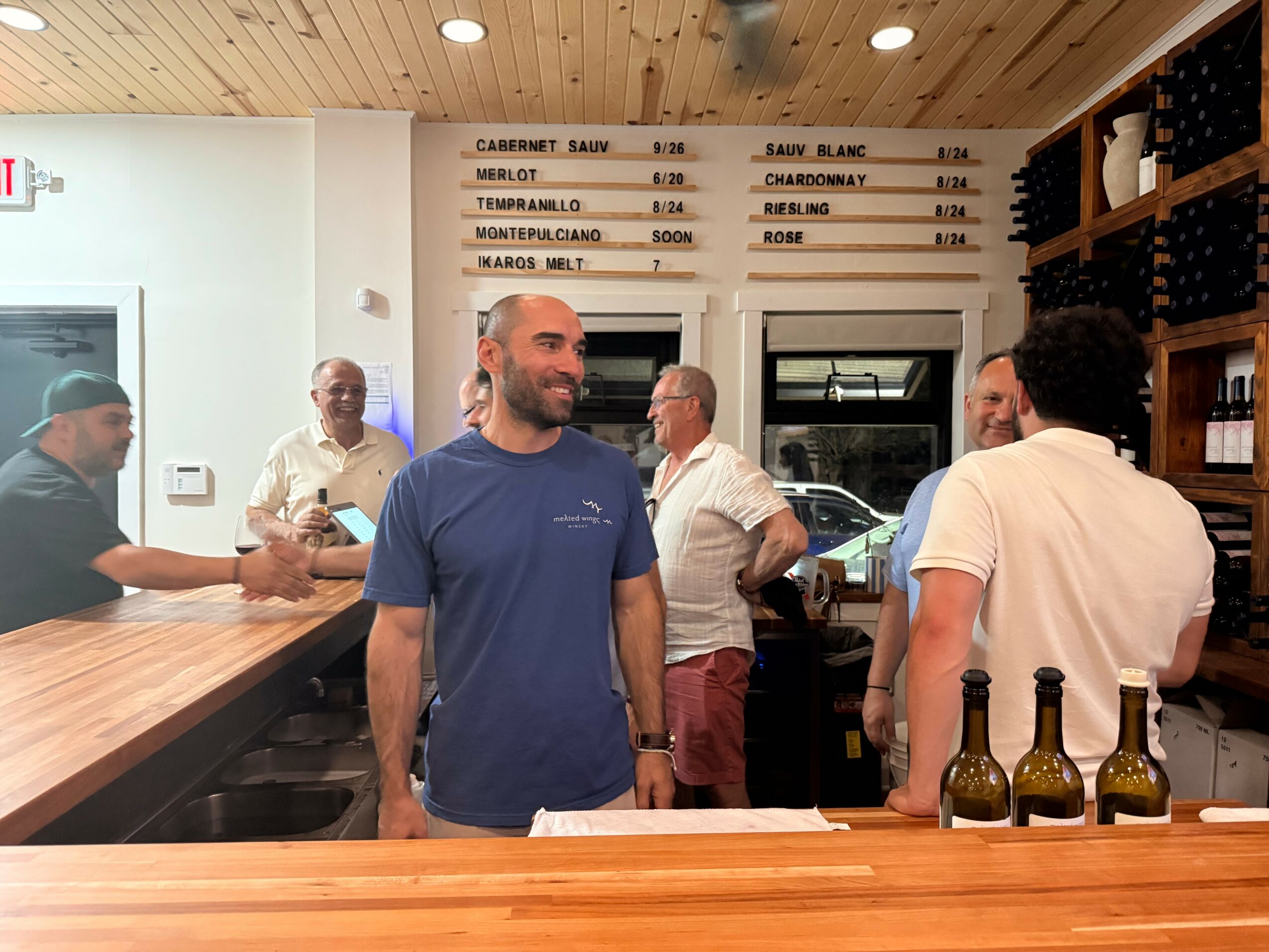 Group of people inside a wine bar, some talking and smiling, with bottles on the counter and a menu board above.
