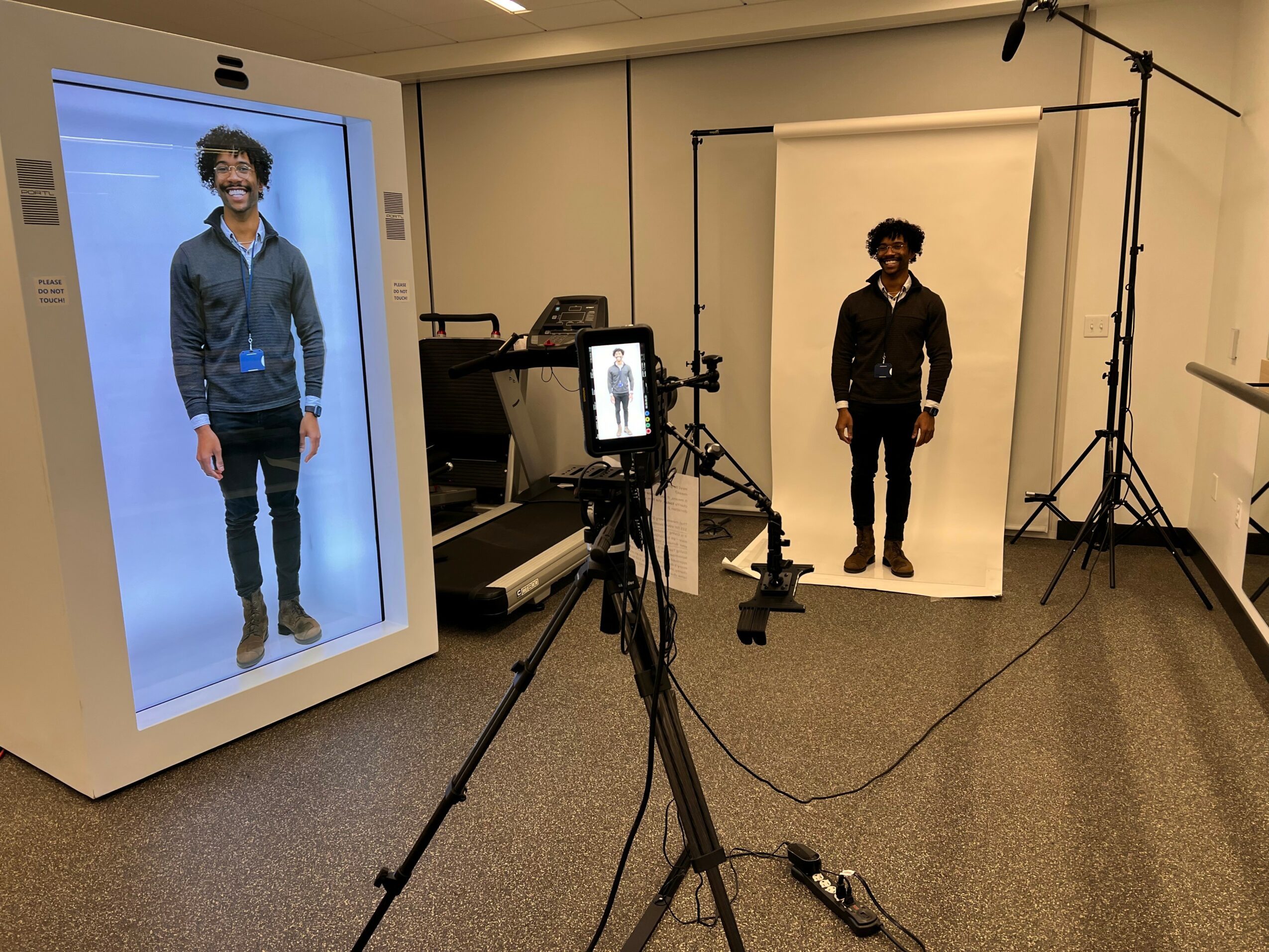 Person standing in a photo studio with a backdrop, camera, and lighting equipment, wearing dark clothing and glasses.