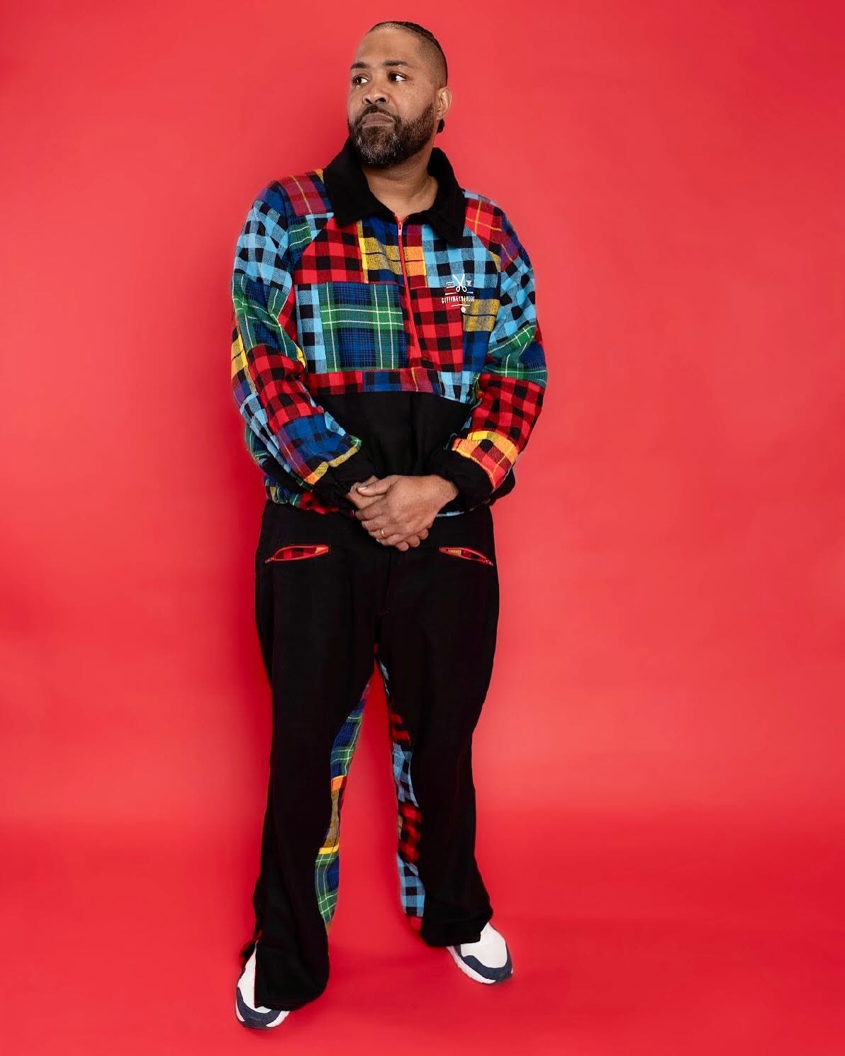 Man with a beard and short hair stands against a red background, wearing a colorful patchwork jacket and black pants.
