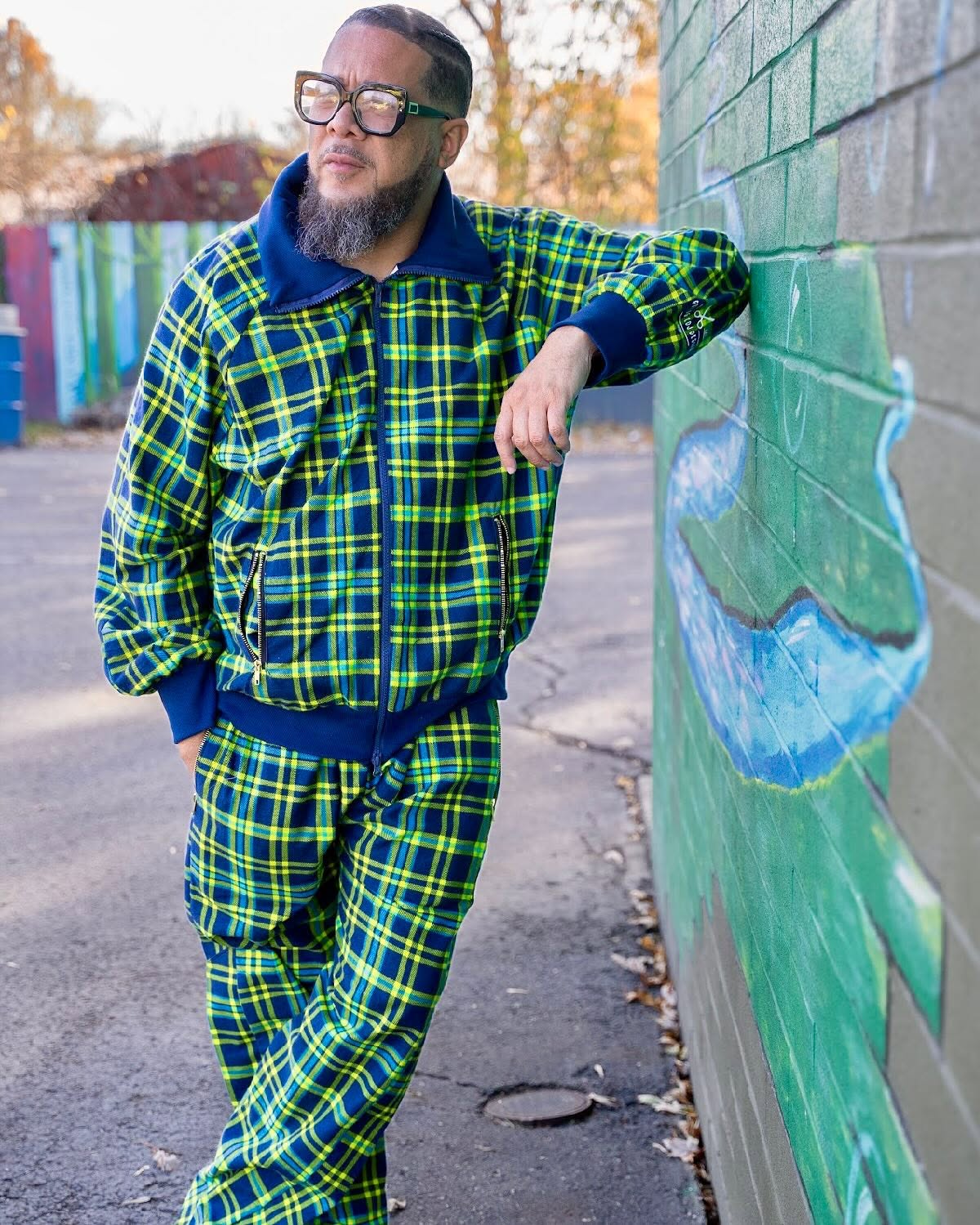 Man with glasses and beard leaning against a wall outdoors, wearing a green and blue plaid outfit, with trees in background.
