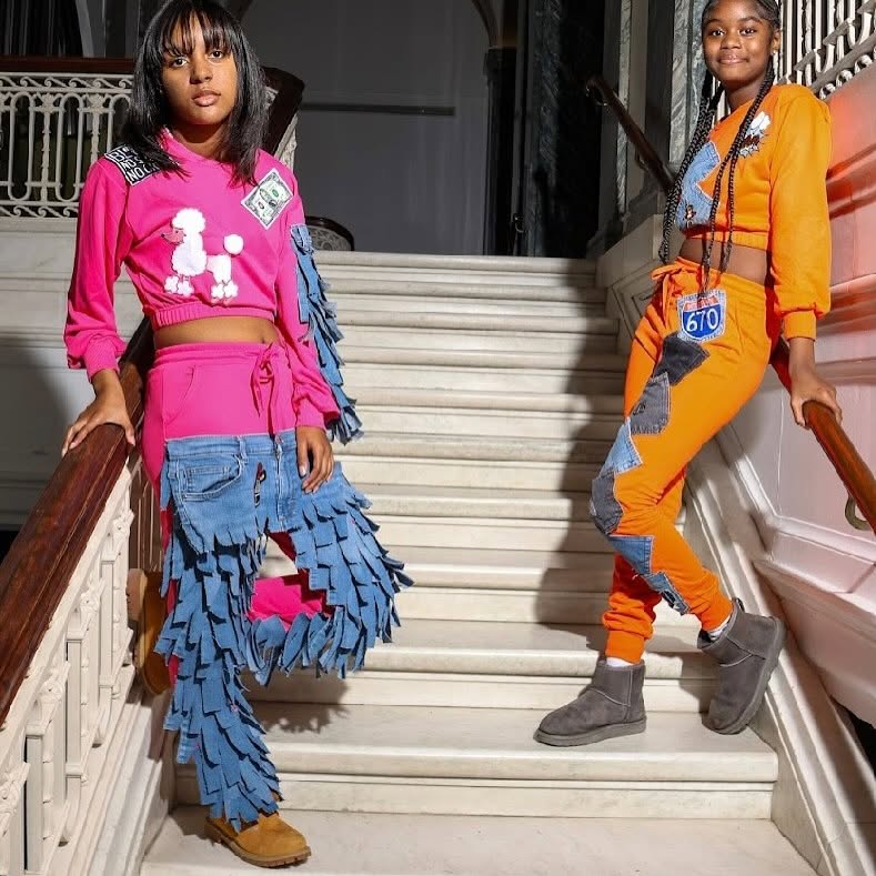 Two young girls pose on a staircase, one in a pink top and fringed skirt, the other in an orange tracksuit, both with badges.