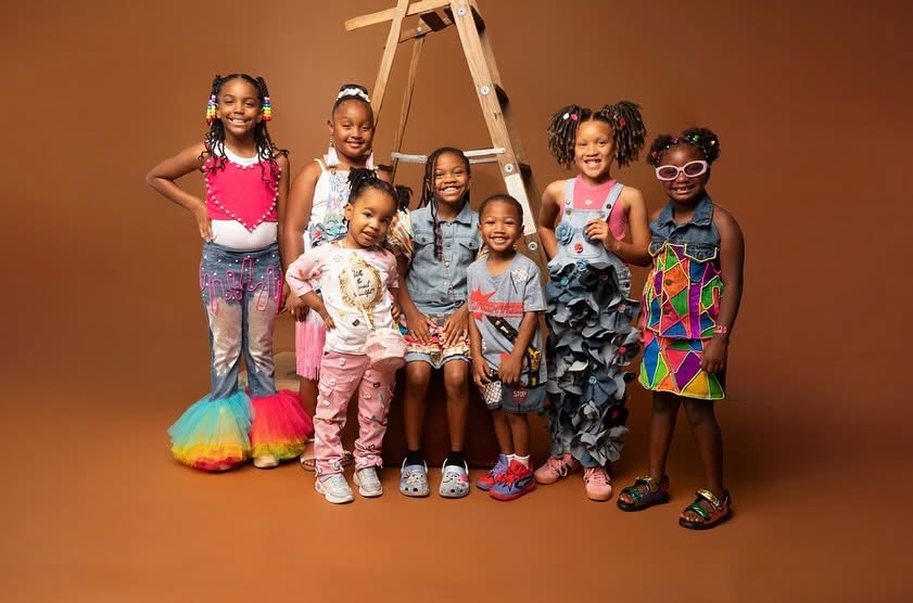 Group of eight children standing together, some smiling, in colorful outfits, with a brown background and a ladder behind them.