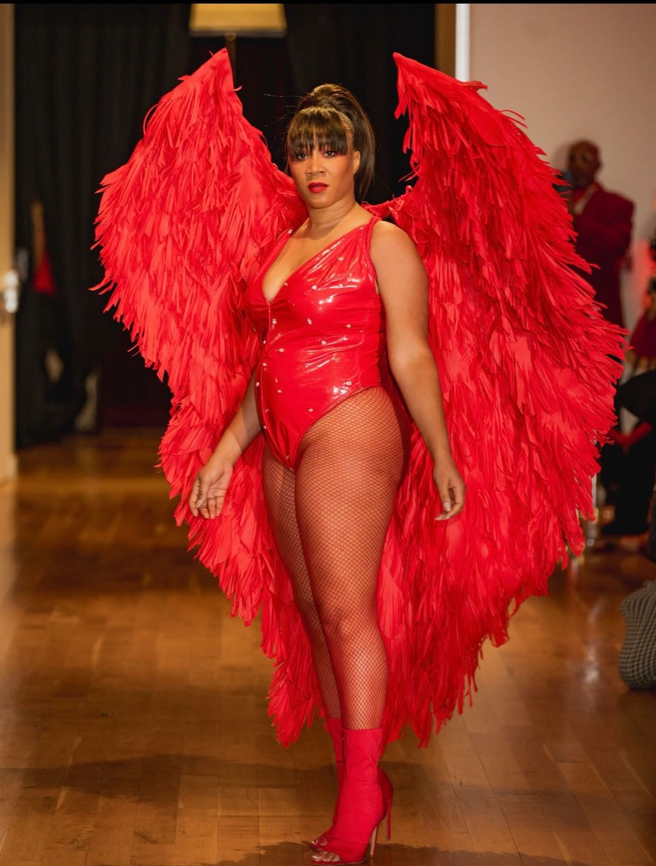Woman in red costume with large feathered wings, fishnet stockings, and matching shoes, standing on a wooden floor indoors.