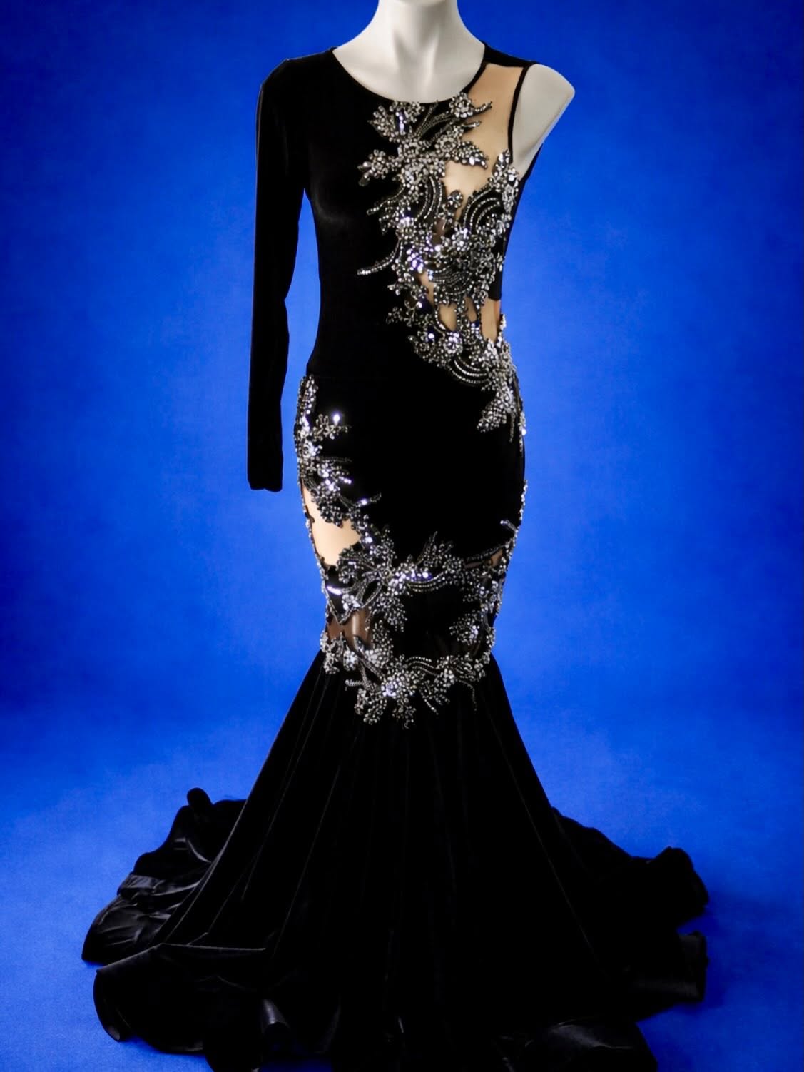 Elegant black gown with intricate silver embroidery and sheer fabric on one shoulder, displayed on a mannequin against a blue background.