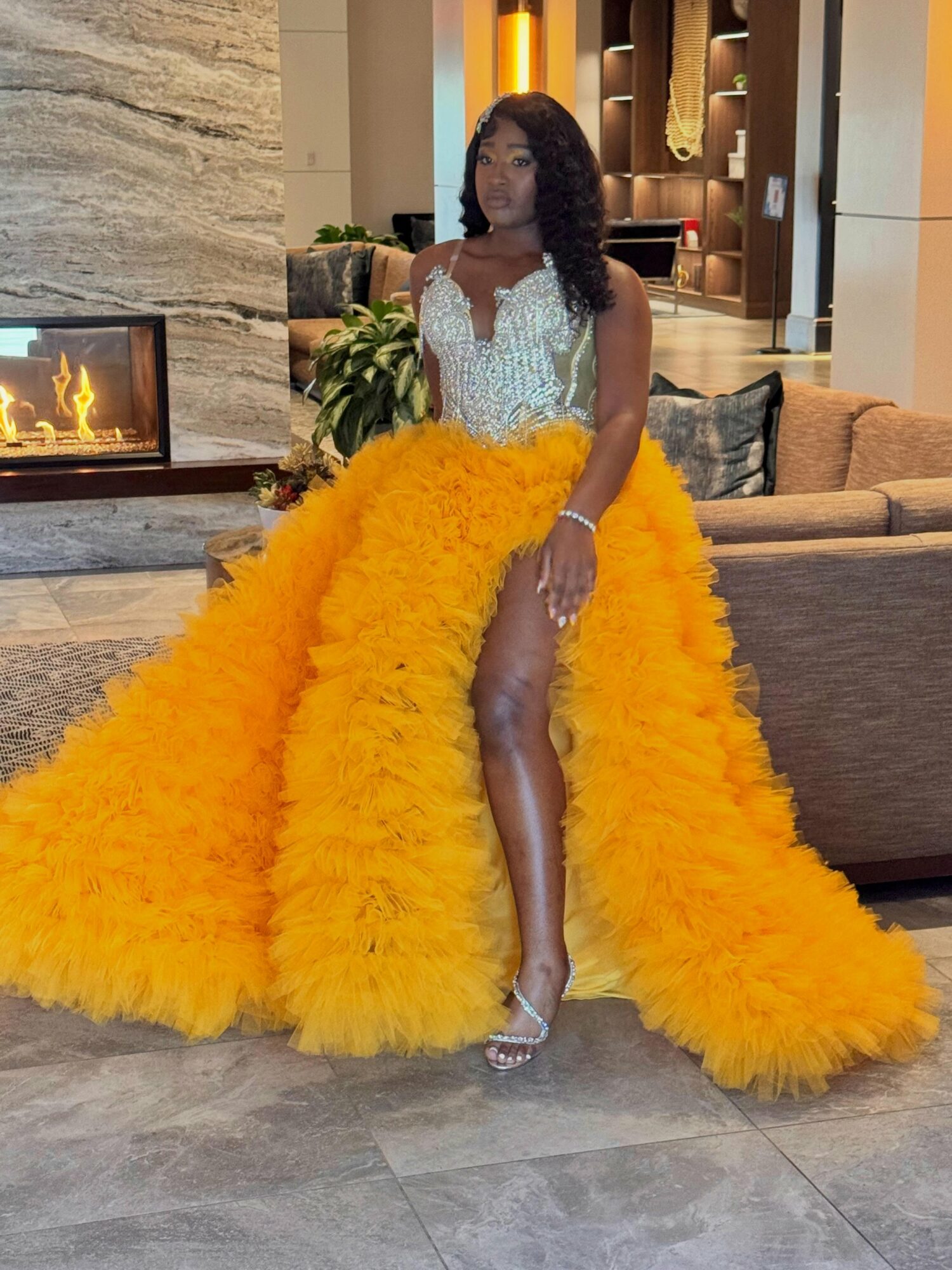 Woman in a yellow feathered gown with a high slit, standing in a modern interior with a fireplace and sofa.