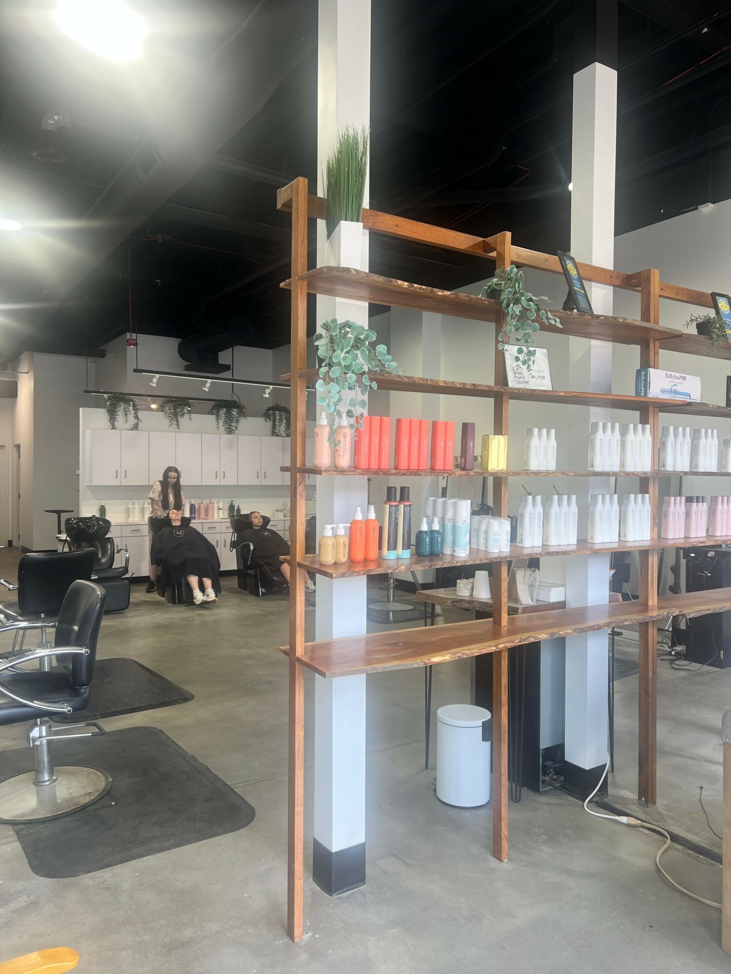 Display shelf with various bottles and plants, people walking in the background, in a store or cafe setting.