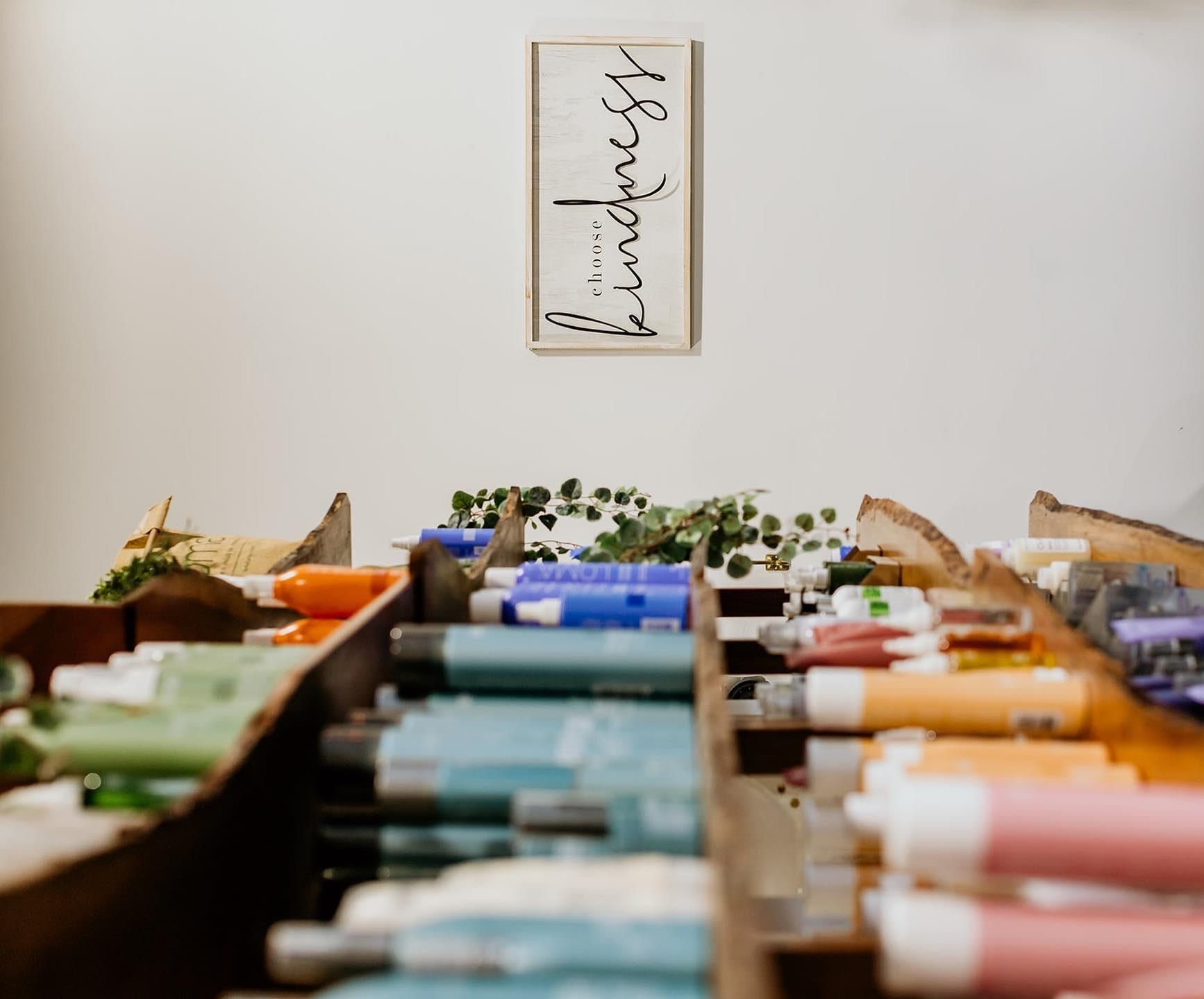 Shelves filled with colorful bottles and containers, with a white wall and a framed sign that reads 'kindness' in cursive.
