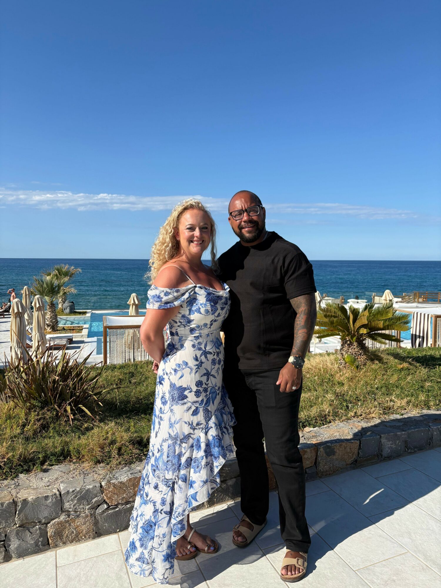 Two women stand outdoors near the ocean, with blue sky and palm trees in the background, smiling at the camera.