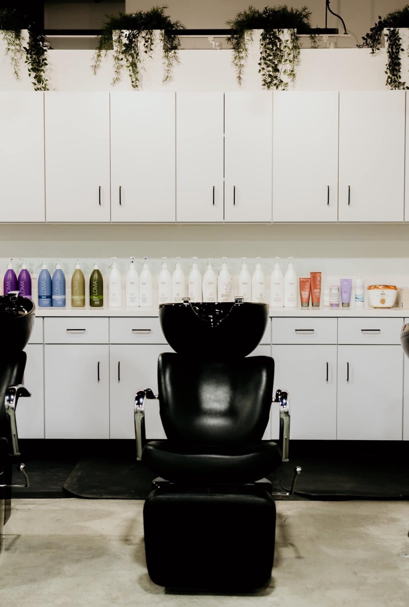 Salon with black chair, white cabinets, bottles, and plants on top, neutral-colored floor, and a black basin.