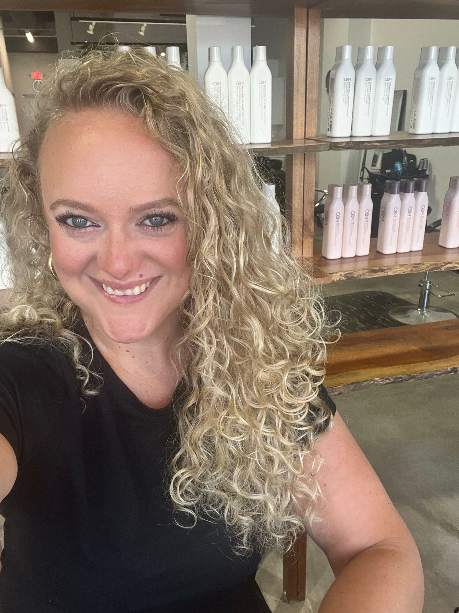 Woman with curly blonde hair smiling, wearing a black top, in a store with shelves of bottles behind her.