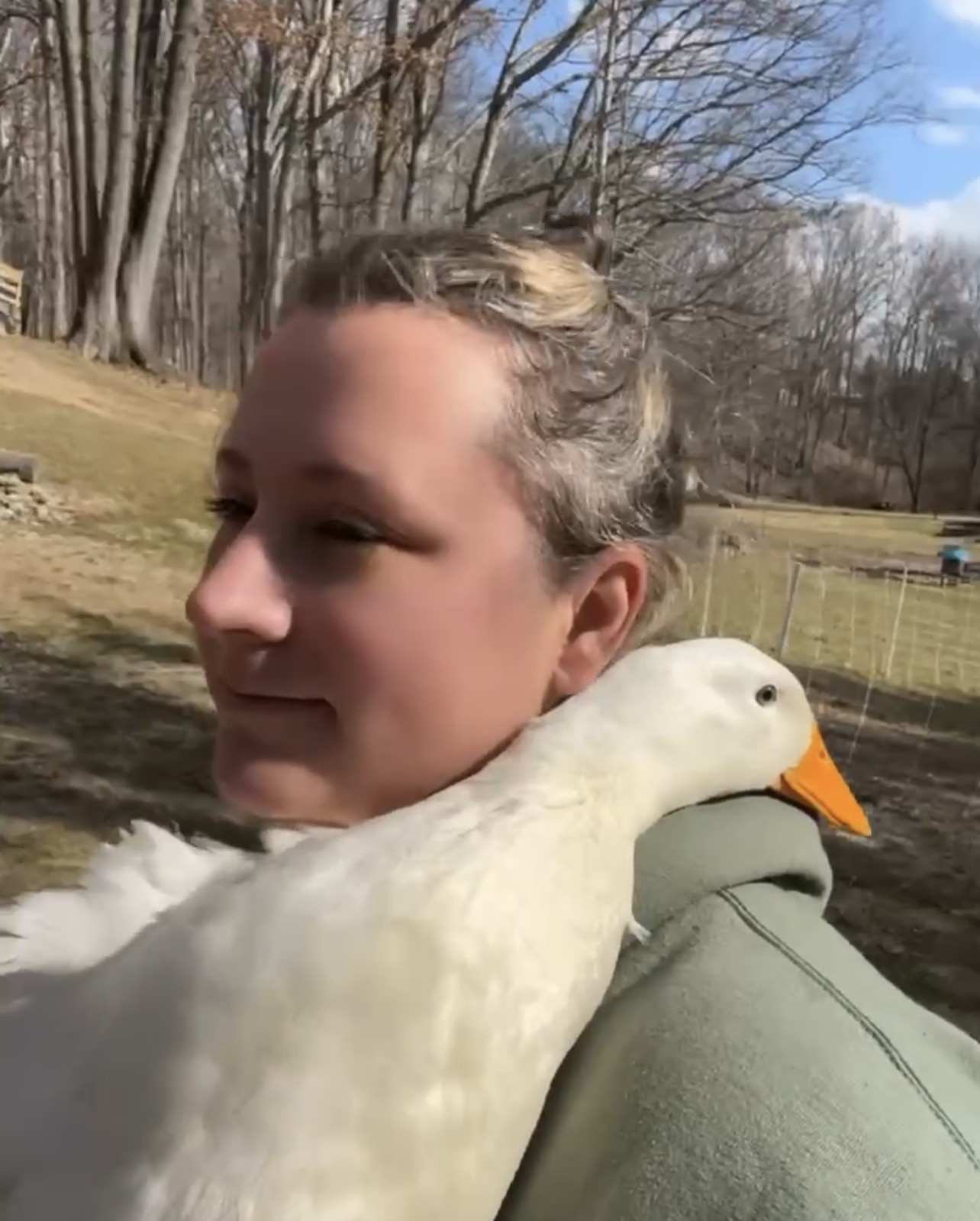 Person with a white duck resting on their shoulder outdoors, trees and a fence in the background.