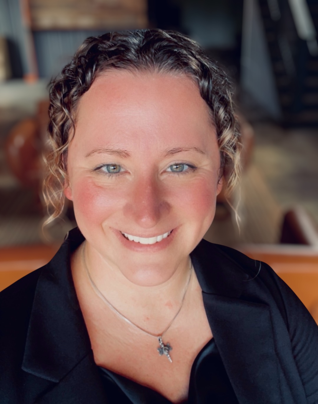 Close-up of smiling woman with curly hair, wearing a black top and a necklace, indoors.