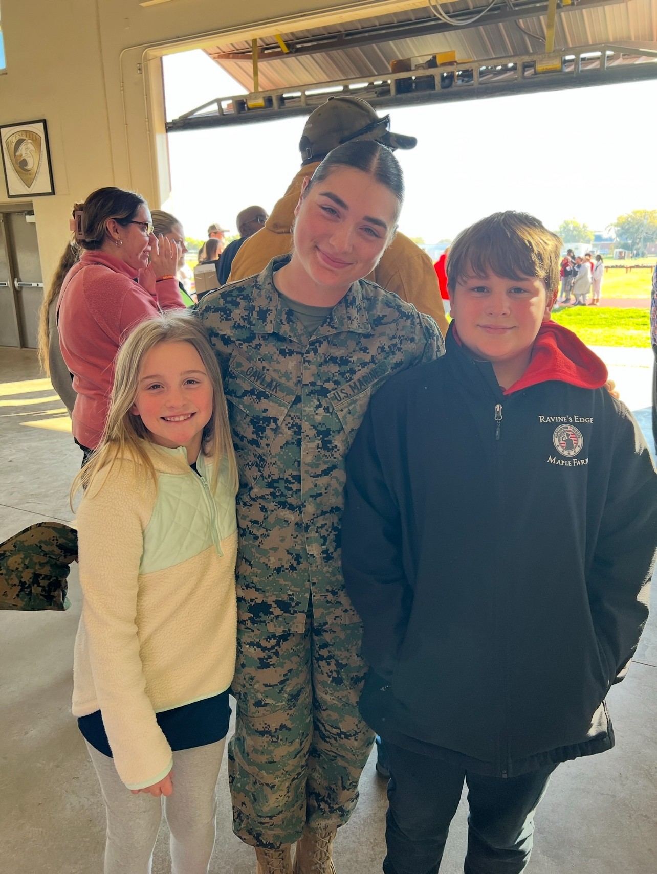 Person in military uniform with two children indoors, other people in background, bright outdoor scene visible through open door.