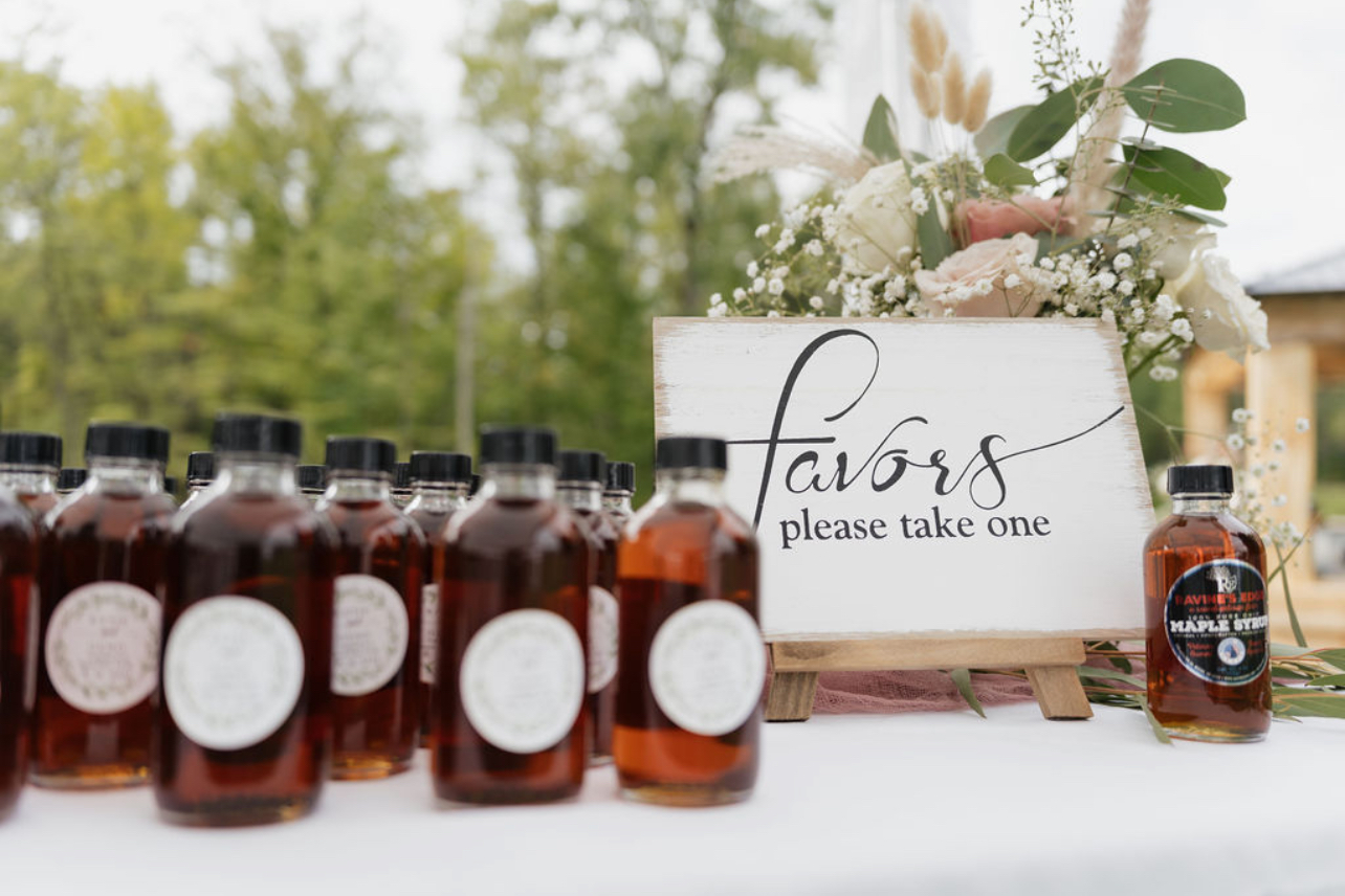 Bottles of honey arranged in front of a floral sign that says 'favor, please take one,' outdoors with trees in background.