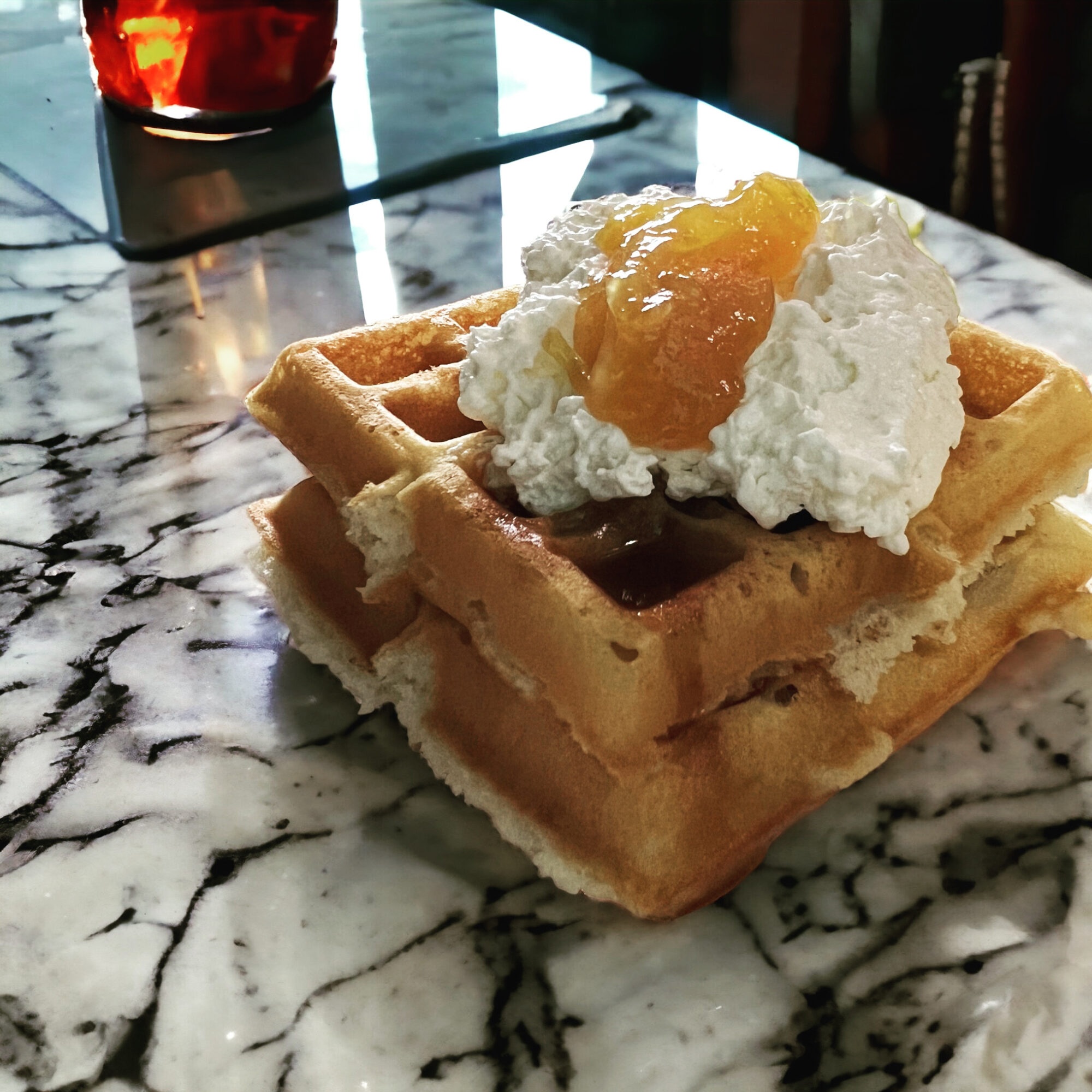 Two waffles topped with whipped cream and fruit preserves on a marble table, with a window and candle in the background.