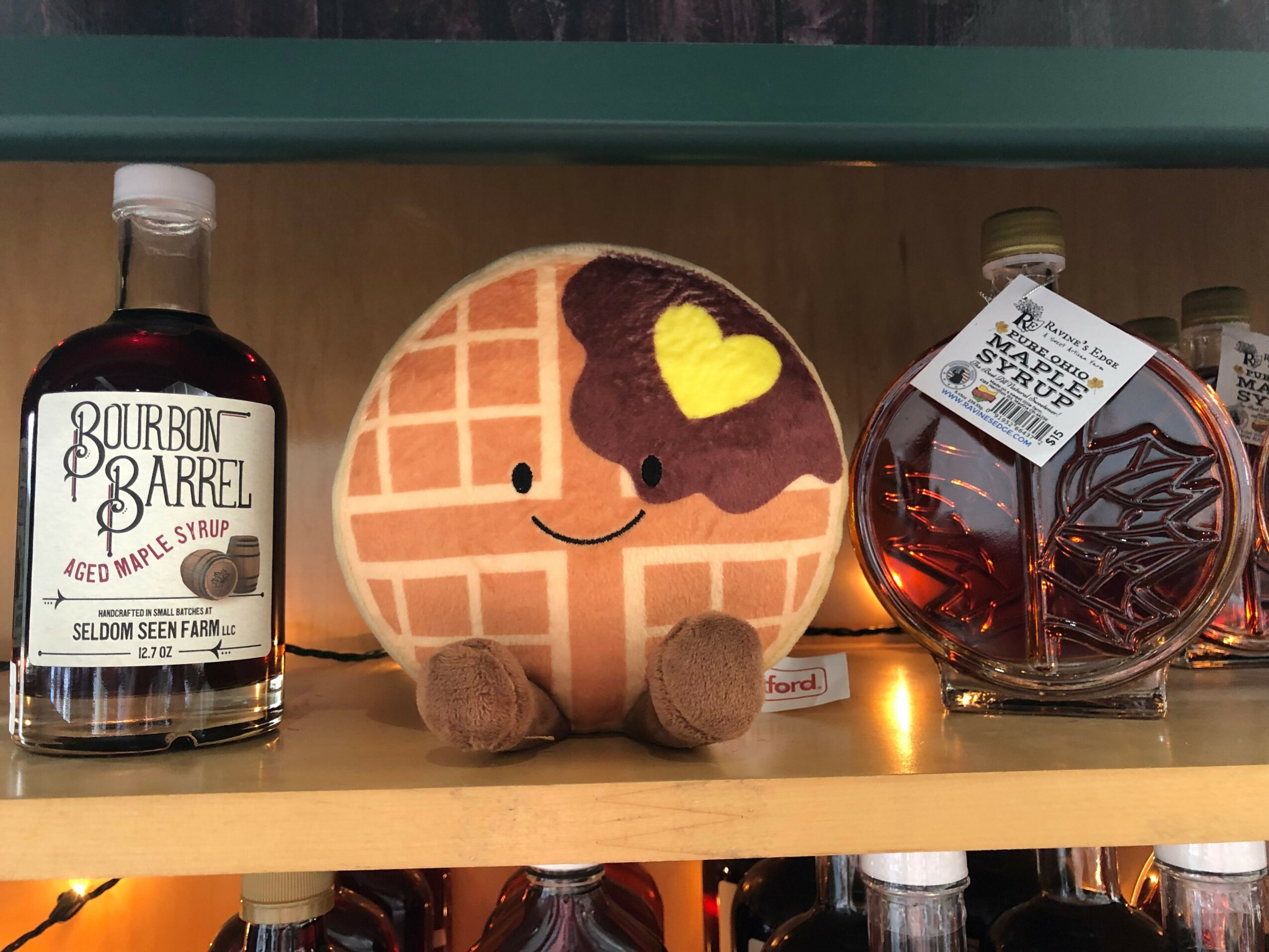 Shelf with a plush waffle character with a heart, bourbon barrel syrup bottle, and a jar of candies.