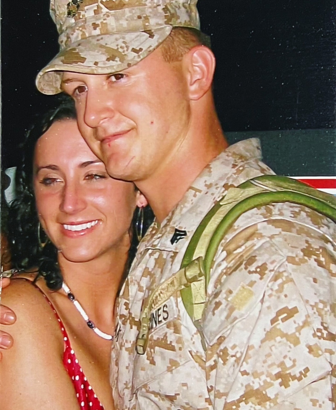 A soldier in camouflage uniform and cap stands close to a smiling woman with dark hair, wearing a red dress and necklace.