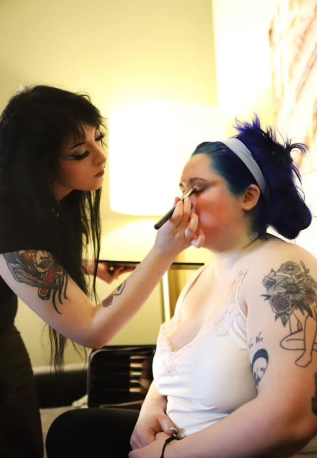 Woman applying makeup to another woman with tattoos, in a room with a lamp and a curtain, both with dark hair.