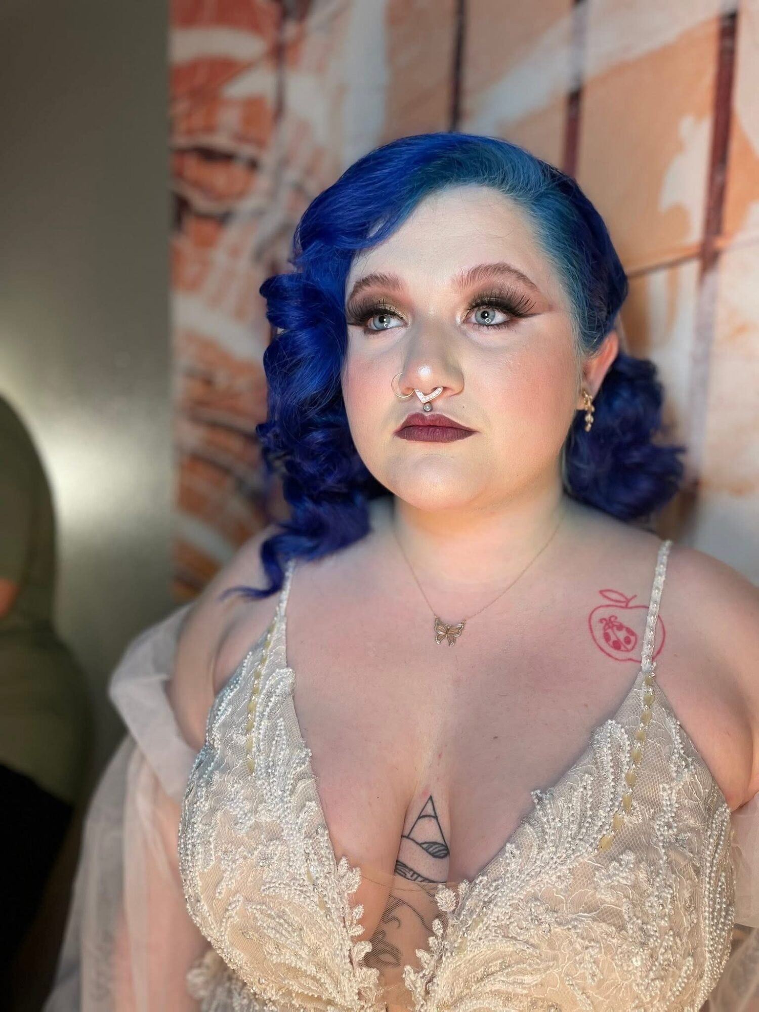 Woman with blue hair, makeup, and a lace top, standing in front of a brick wall.