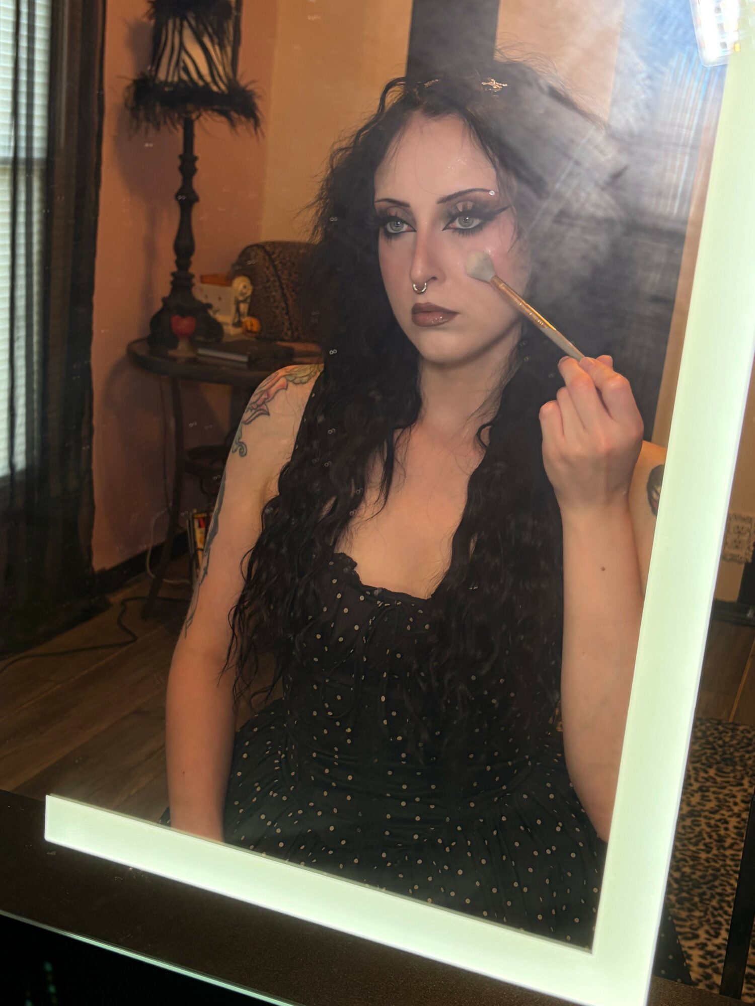 Woman with long dark hair and makeup, wearing a black dress, holding a makeup brush near her face, in a room with warm lighting.