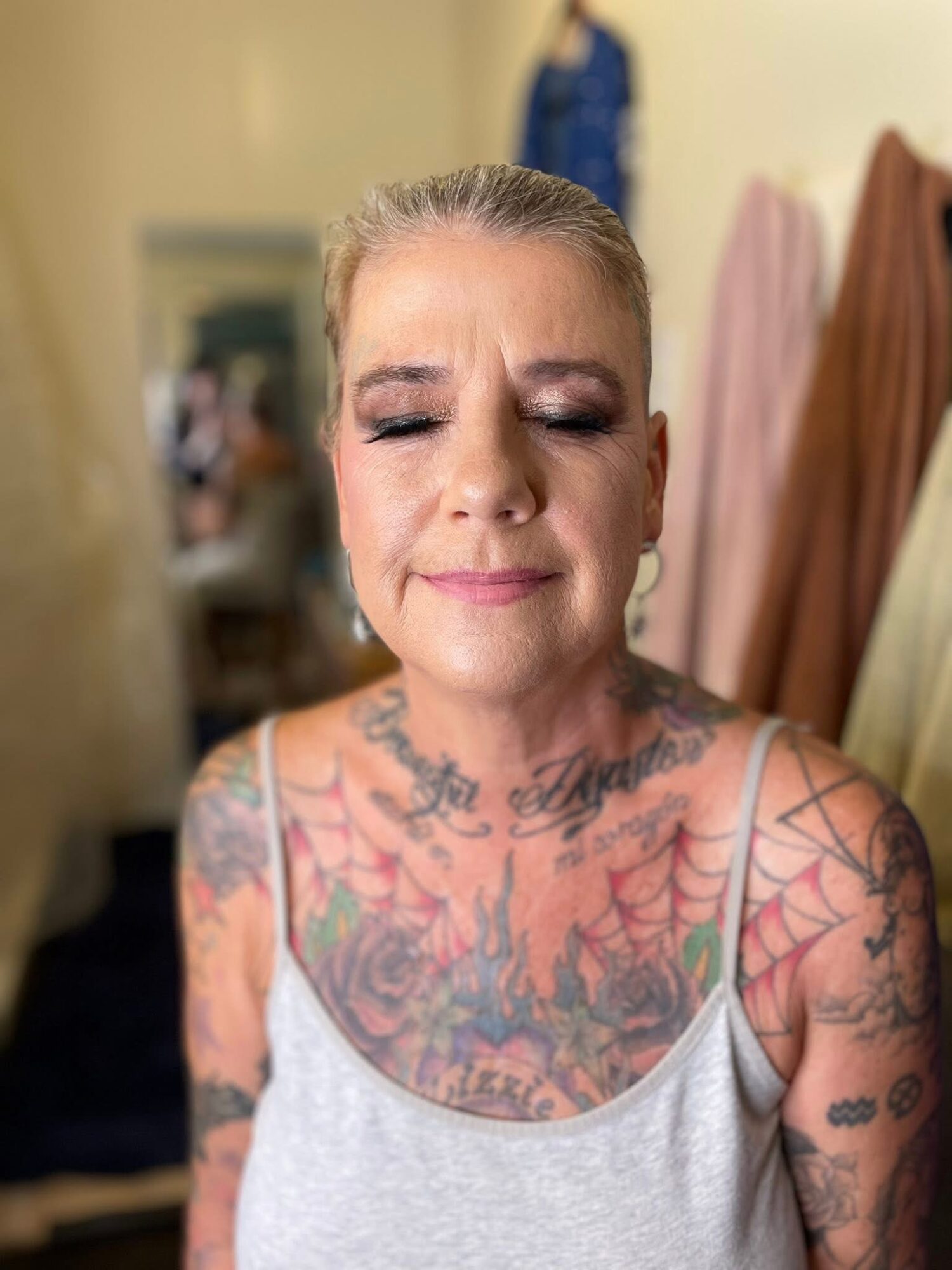 A woman with tattoos on her chest and arms, wearing a light gray tank top, with her eyes closed and a slight smile.