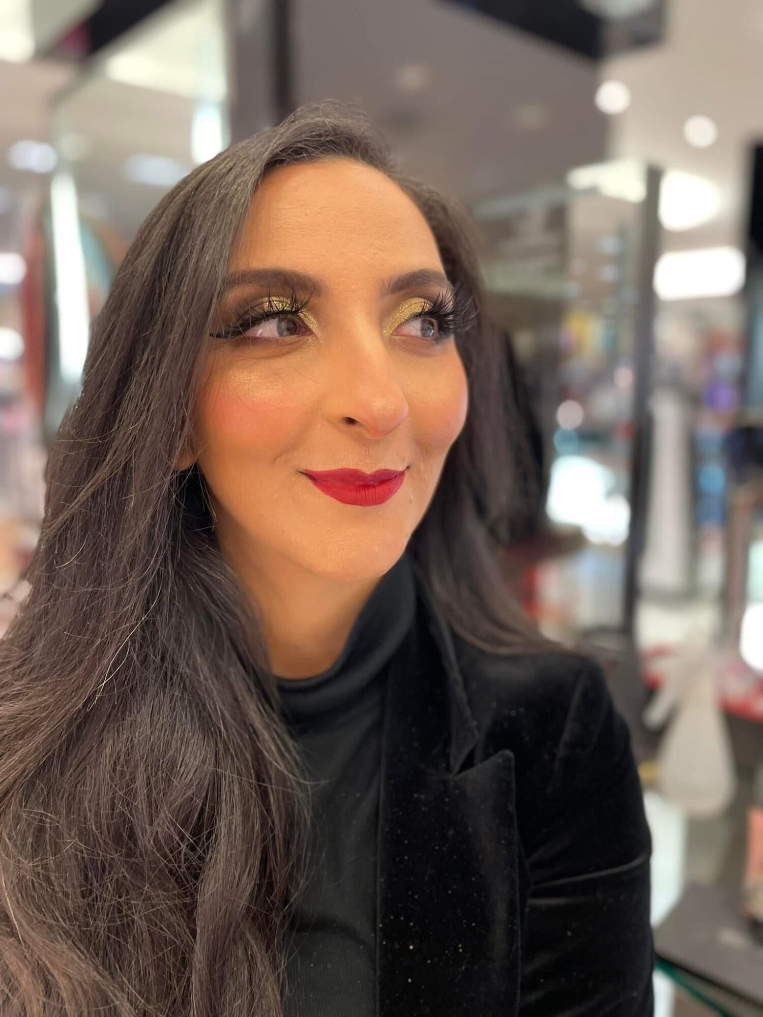 Woman with long dark hair, makeup, and red lipstick, looking to the side in an indoor setting.