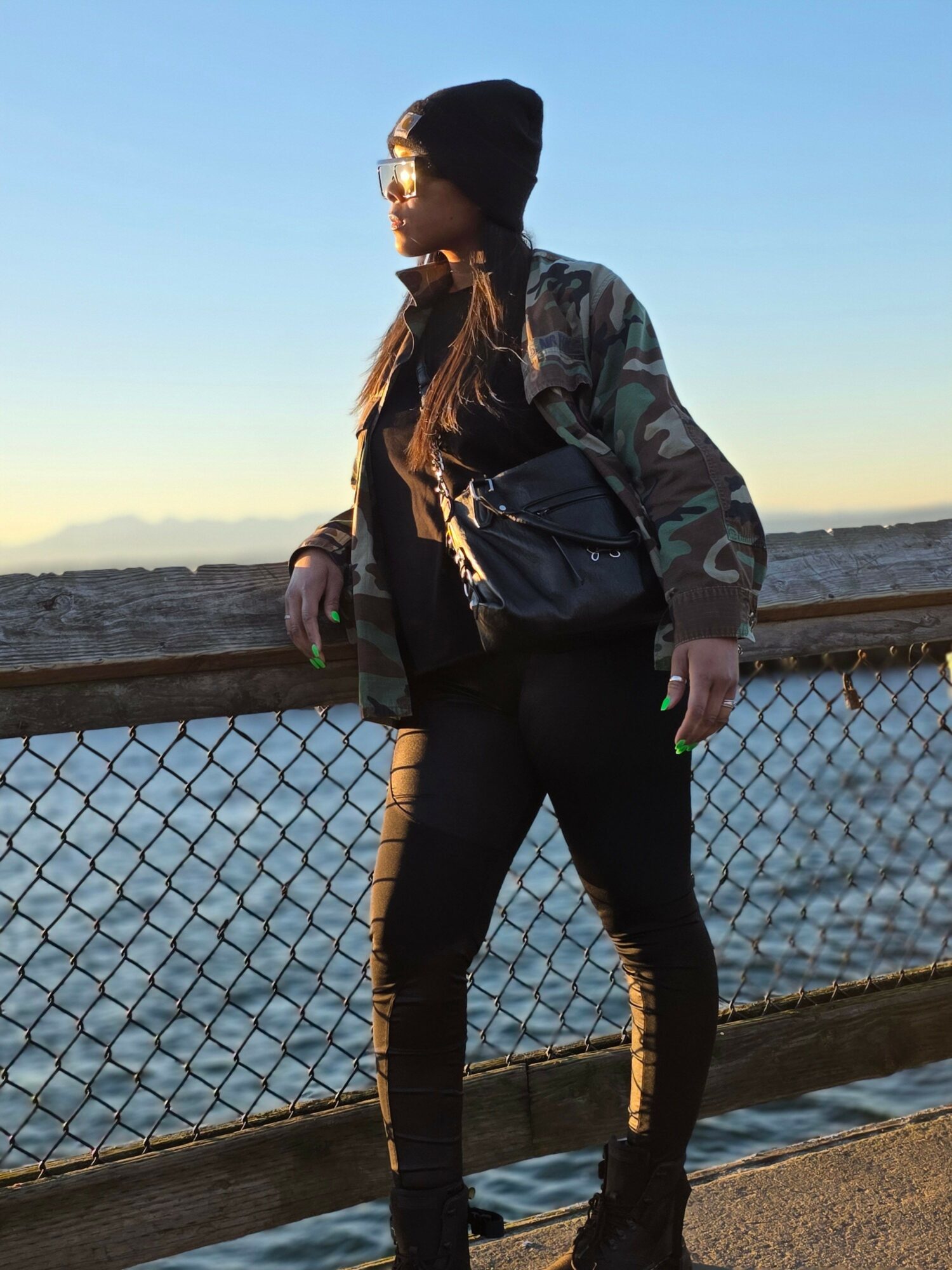 Person standing near a chain-link fence at sunset, wearing a jacket and jeans, with water and sky in the background.