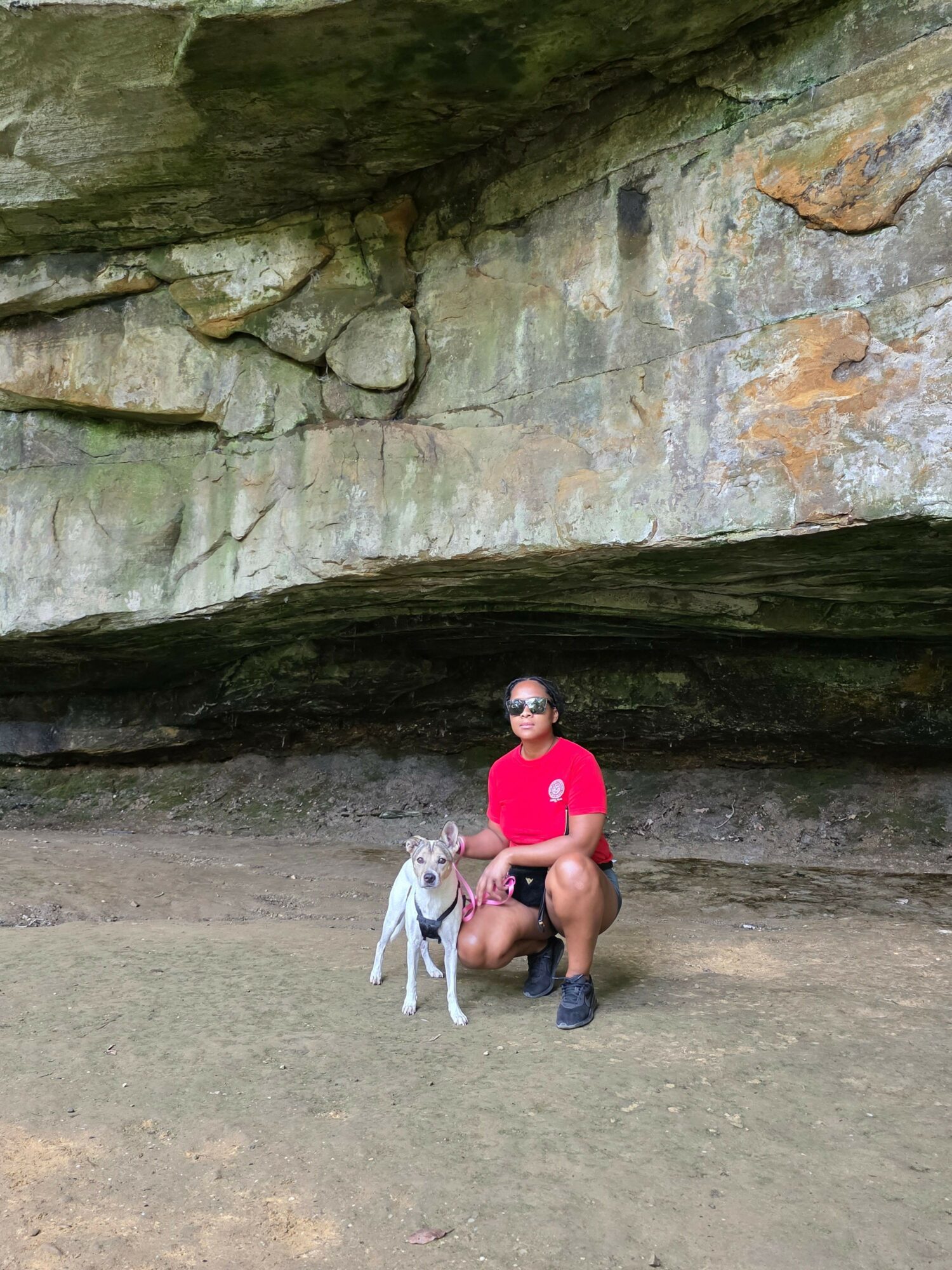 Person sitting on the ground near a stone wall, holding a pink object, with a dog nearby, outdoors.