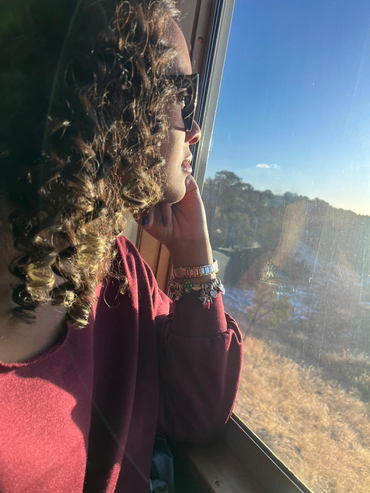 Person with curly hair wearing glasses and a maroon top looking out a window at a landscape with trees and blue sky.