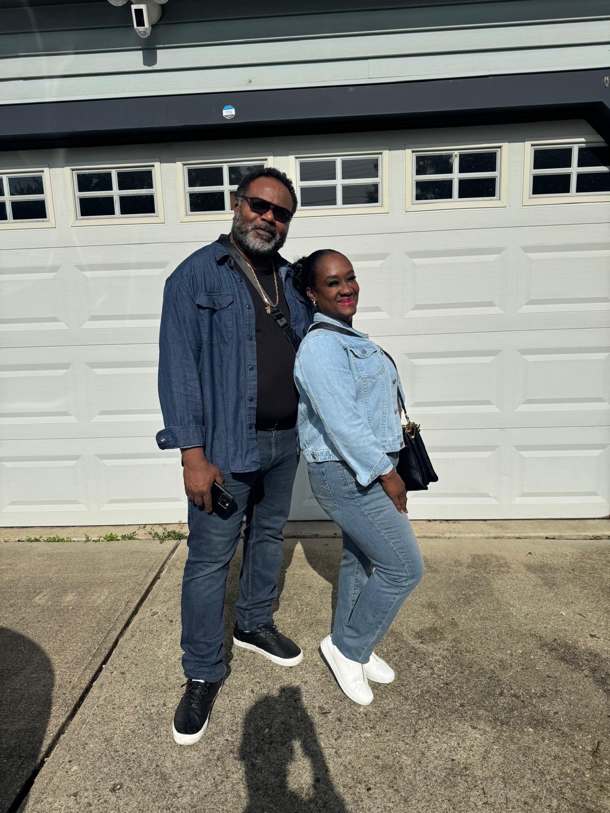 Man and woman standing outside in front of a garage door, smiling, with shadows on the ground.