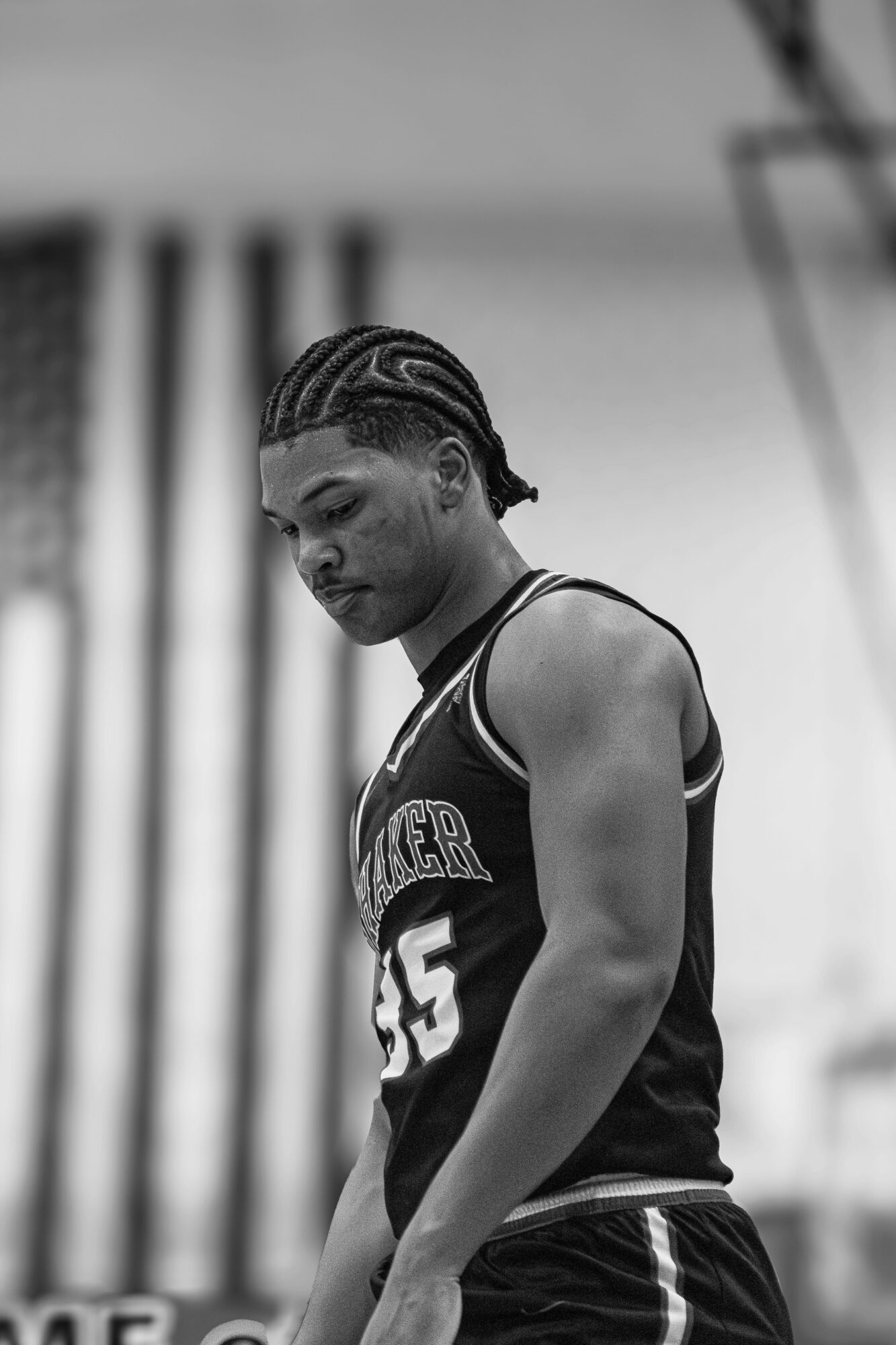 Young female athlete with braided hair, wearing a sports jersey, looking down thoughtfully, outdoors with blurred background.