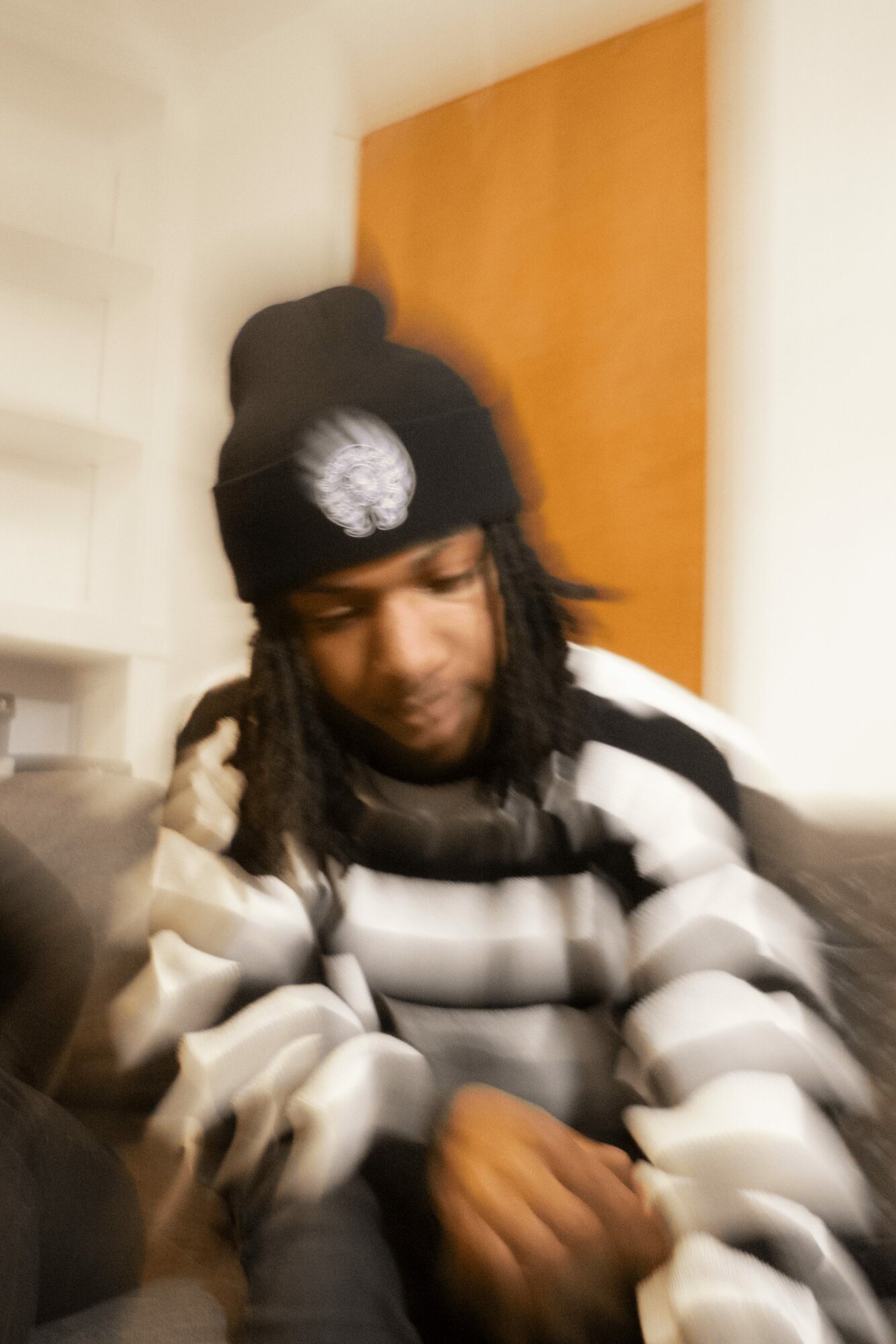 Person wearing a black beanie with a white logo, sitting indoors with a striped jacket, looking down.