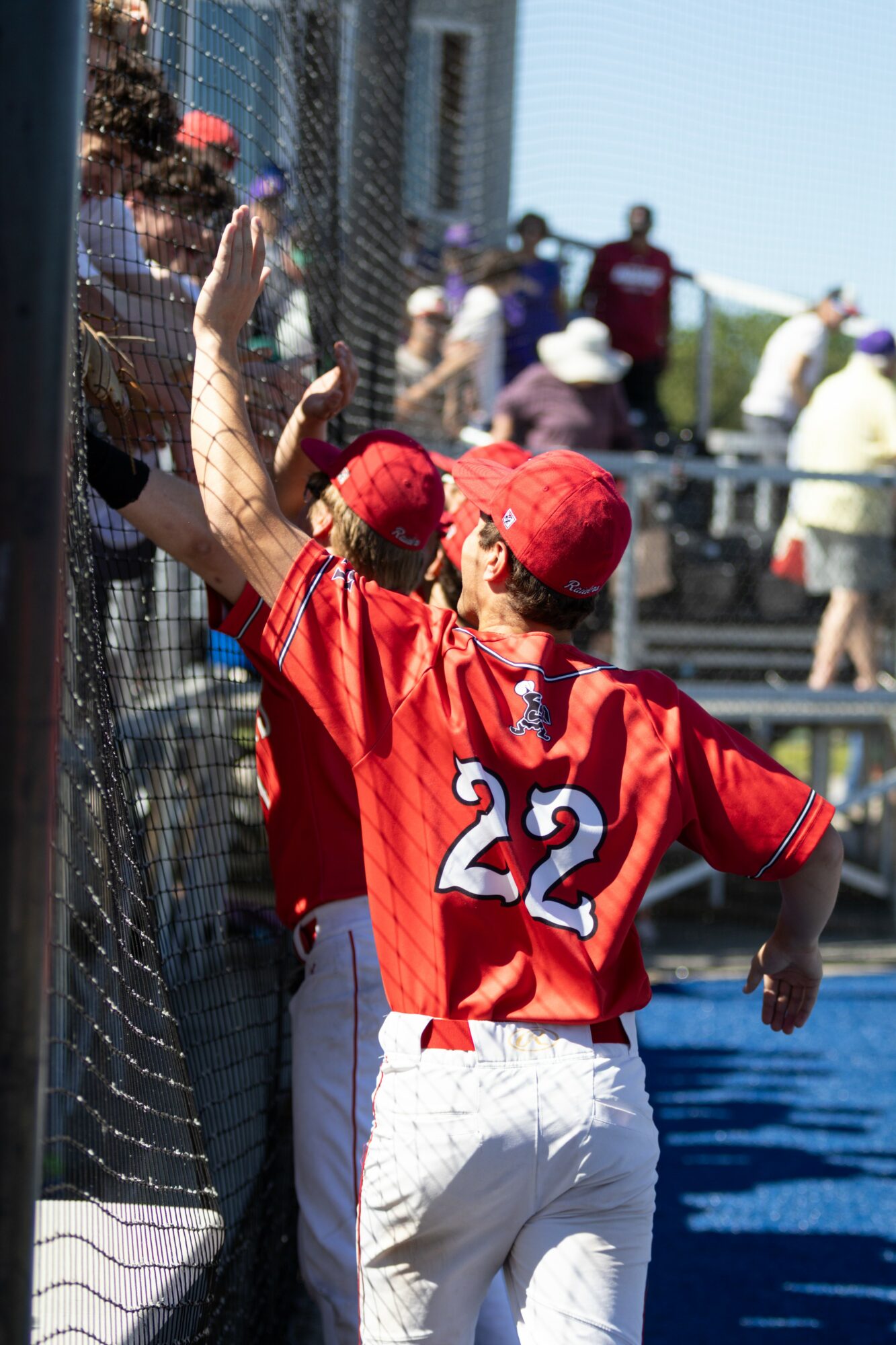Baseball player in red jersey and cap high-fives a person behind a fence, with spectators in background.