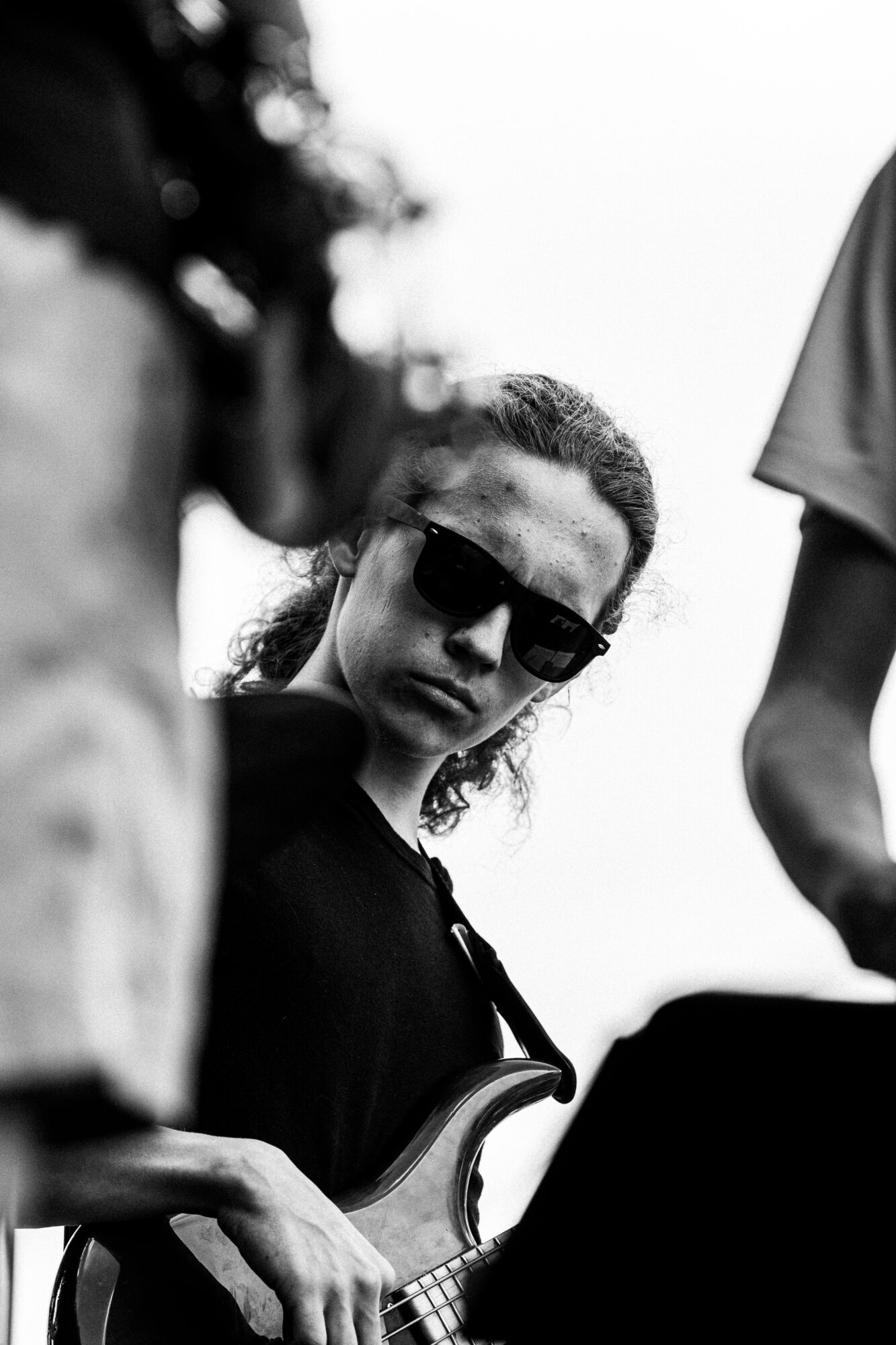 Woman wearing sunglasses holding a guitar, surrounded by people, outdoors, black and white photo.
